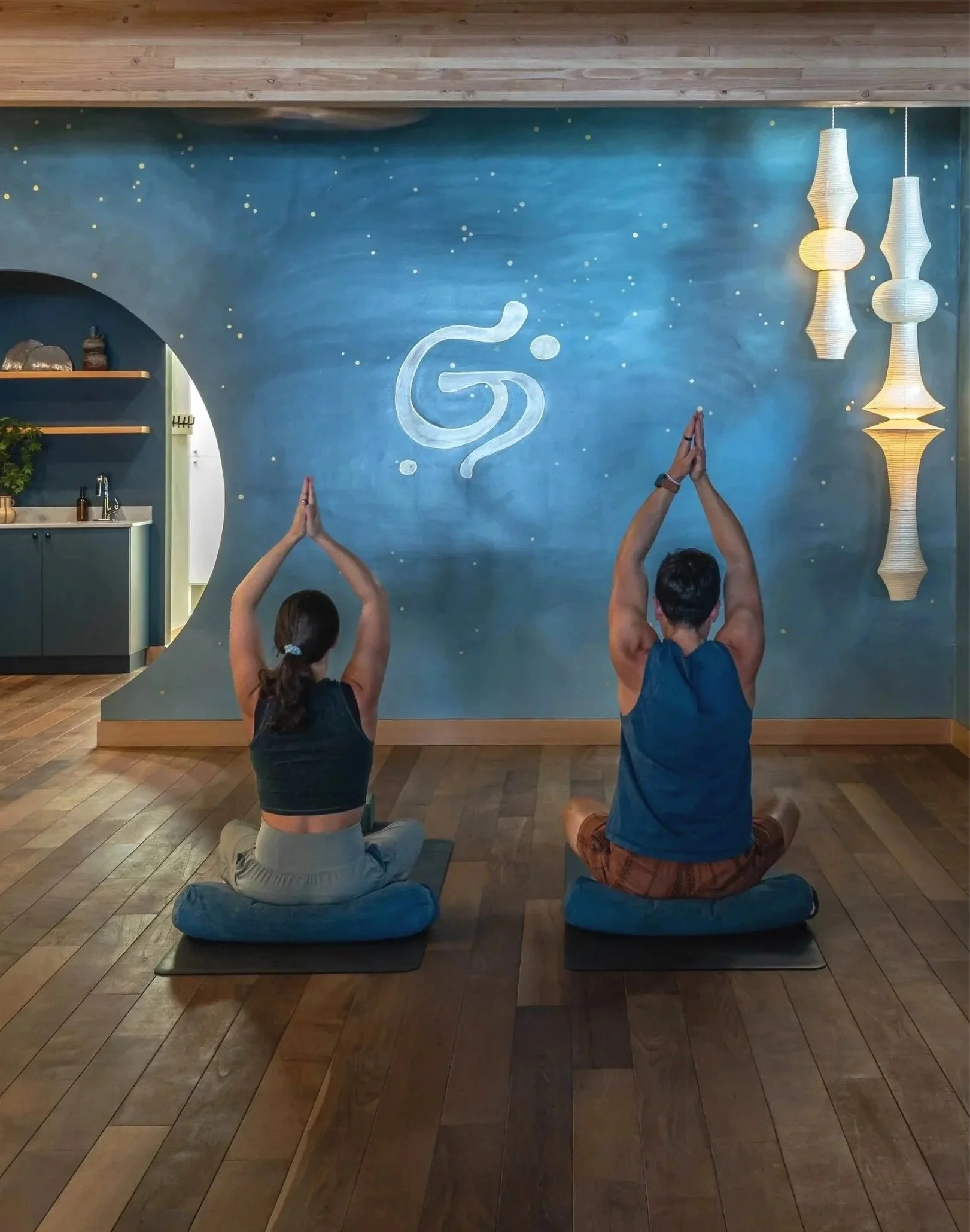 Two people practicing yoga in a yoga studio, sitting on black mats with legs crossed and hands in prayer position, facing a blue wall with a white abstract design and small yellow dots, hanging pendant lights on the right, and kitchen space visible through a circular cutout on the left.