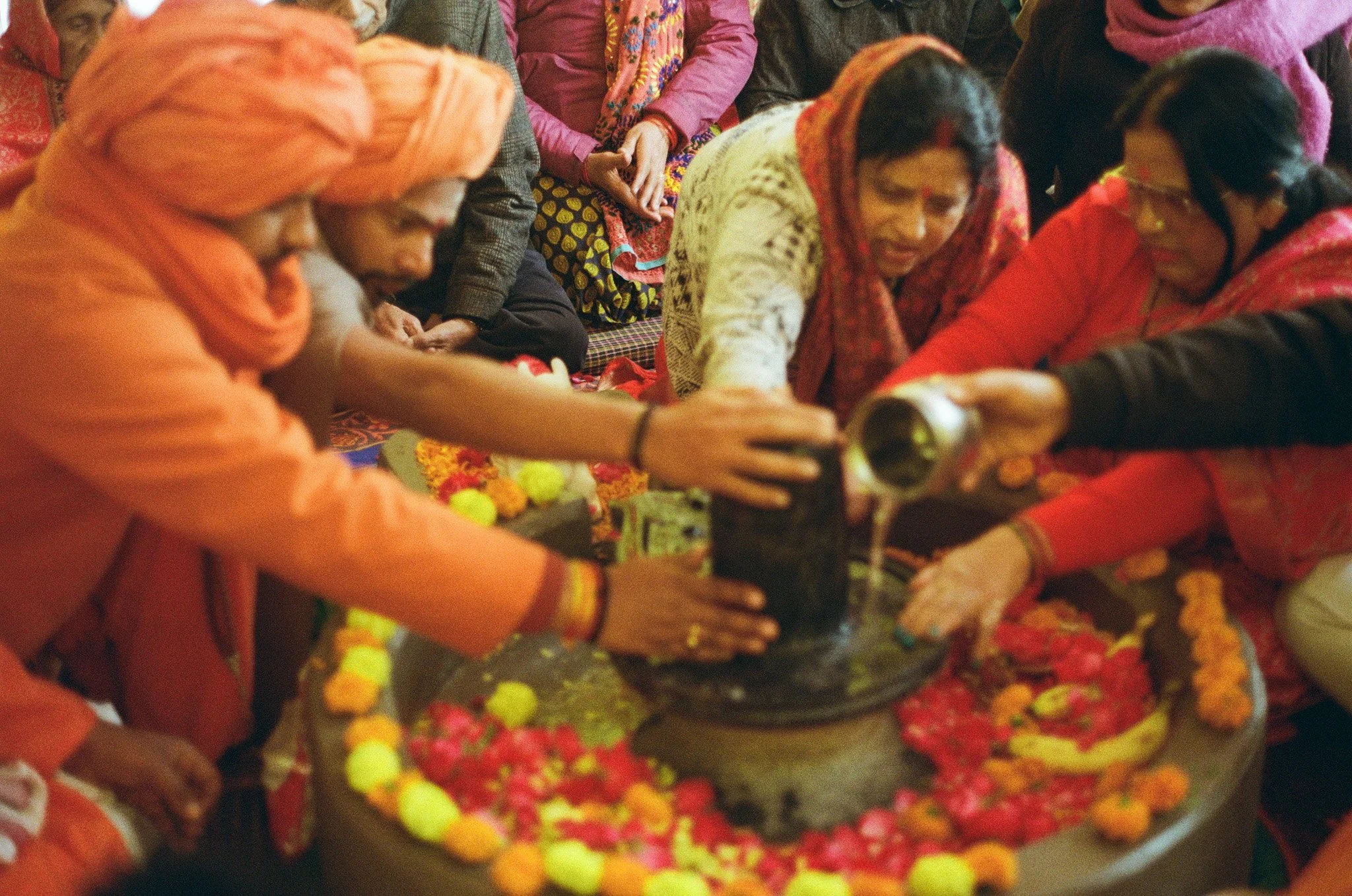 Hindu women performing a traditional ritual with water poured over a Shiva lingam, surrounded by marigold flowers, during a religious ceremony.