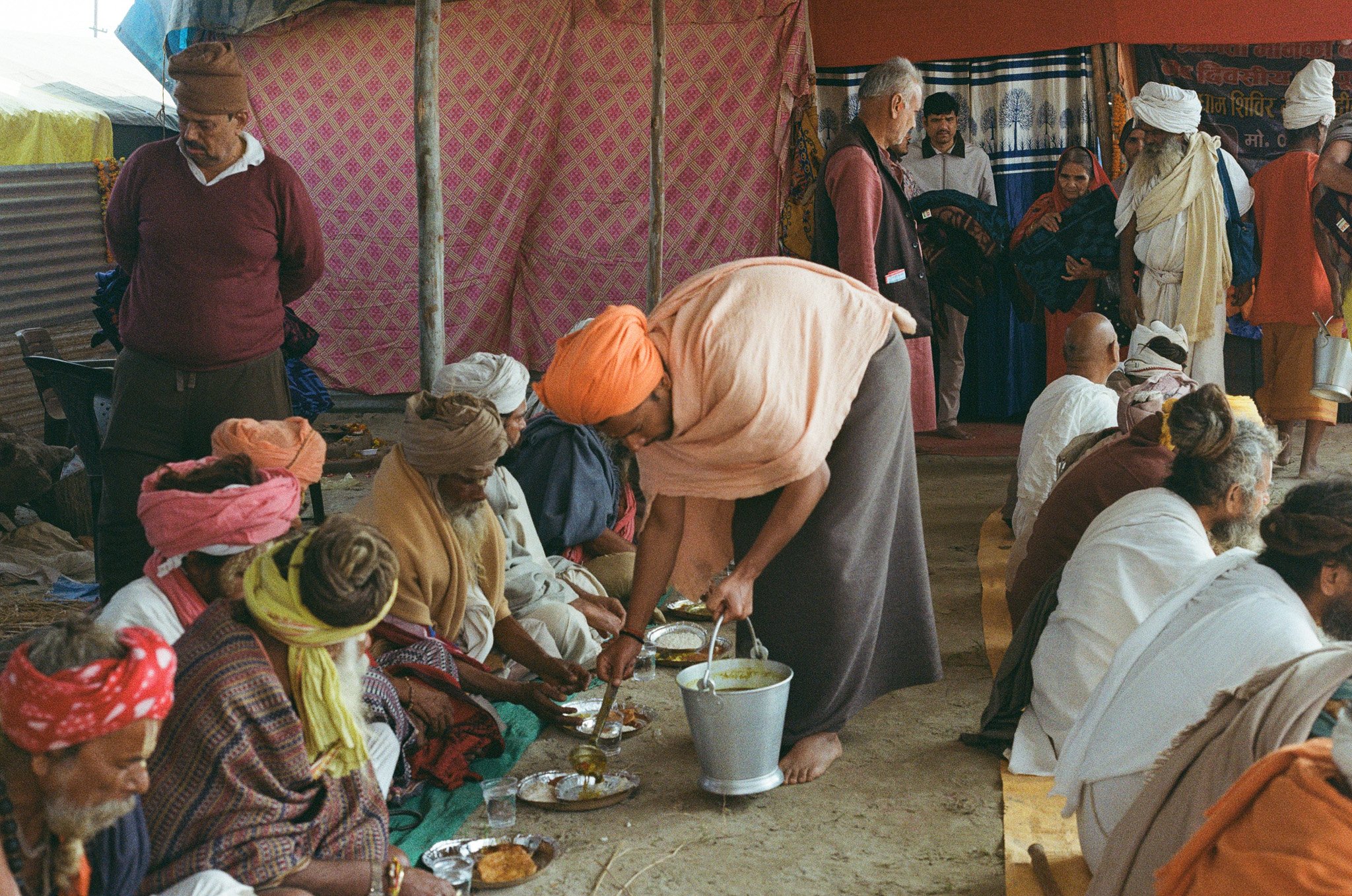 People sitting and standing in a large outdoor tent, some receiving food from a man with a large spoon and bucket, many wearing traditional attire and head coverings, with colorful fabric walls in the background.