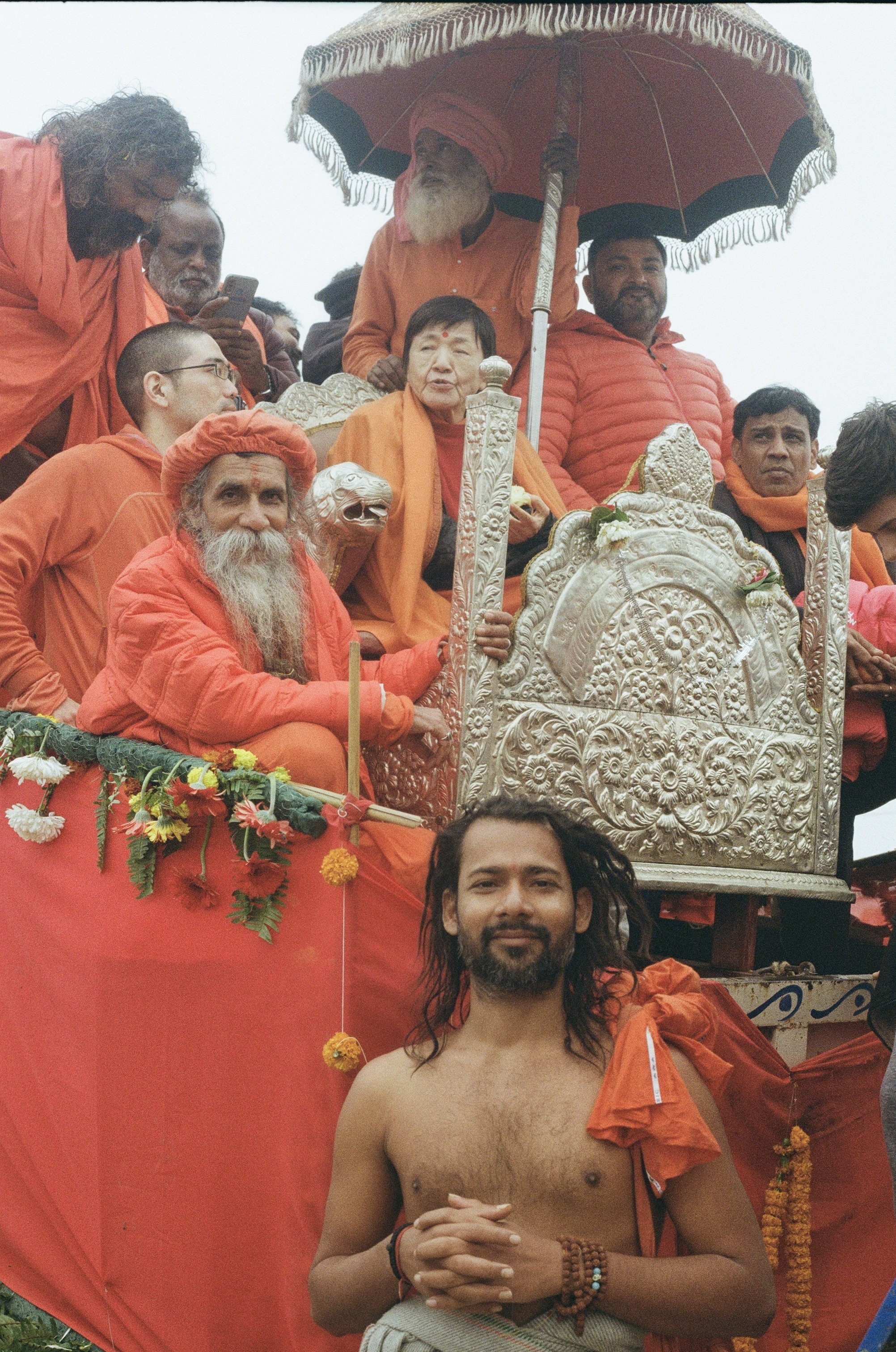 A religious procession with people dressed in orange, including a holy man with a long beard and an orange turban, riding on a decorated chariot. An elderly woman sits beside him, holding a silver staff. A shirtless man with long hair and prayer bead