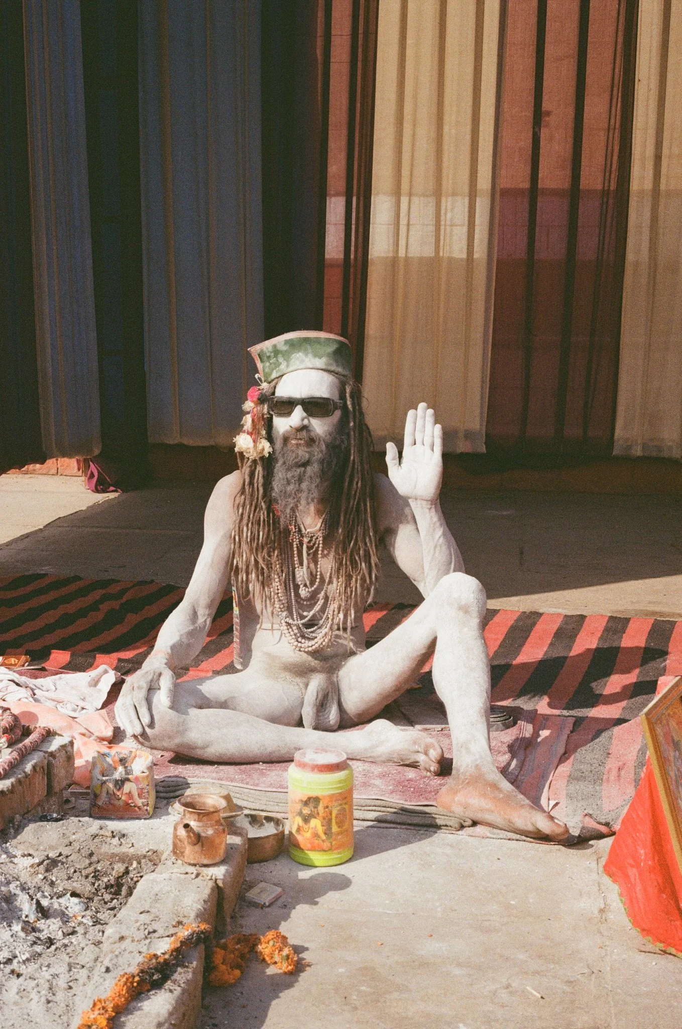 A person with dreadlocks, sunglasses, and a hat sitting cross-legged on a mat, with jewelry and raising one hand in a gesture, in front of a colorful curtain.