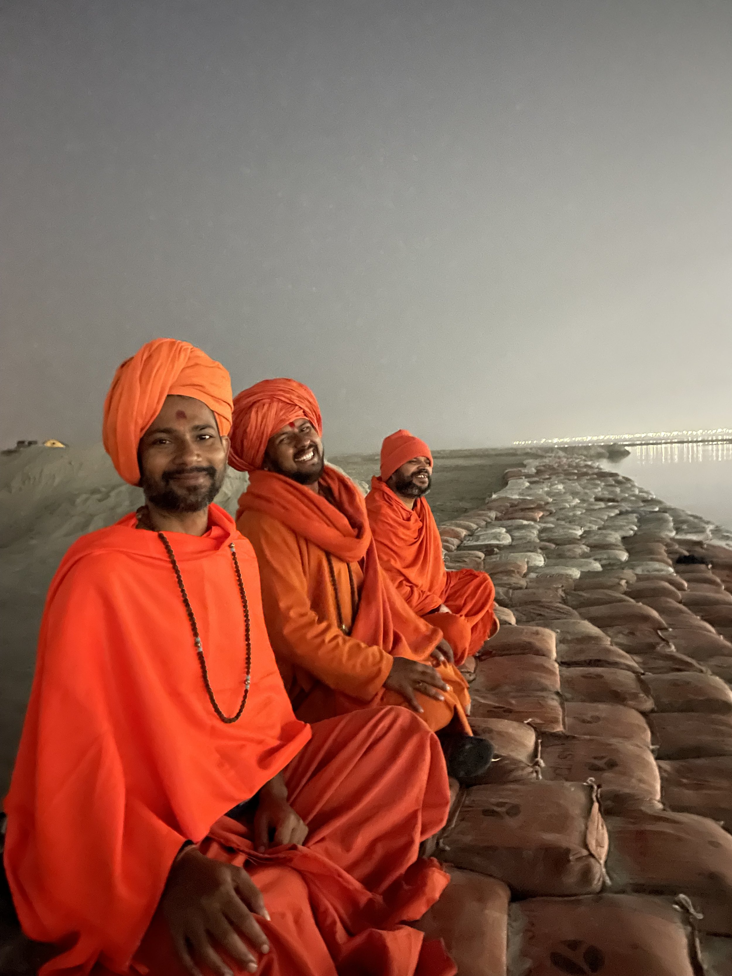 Three Indian men dressed as monks in orange robes sitting on a stone pathway near water at night, with city lights in the distance.
