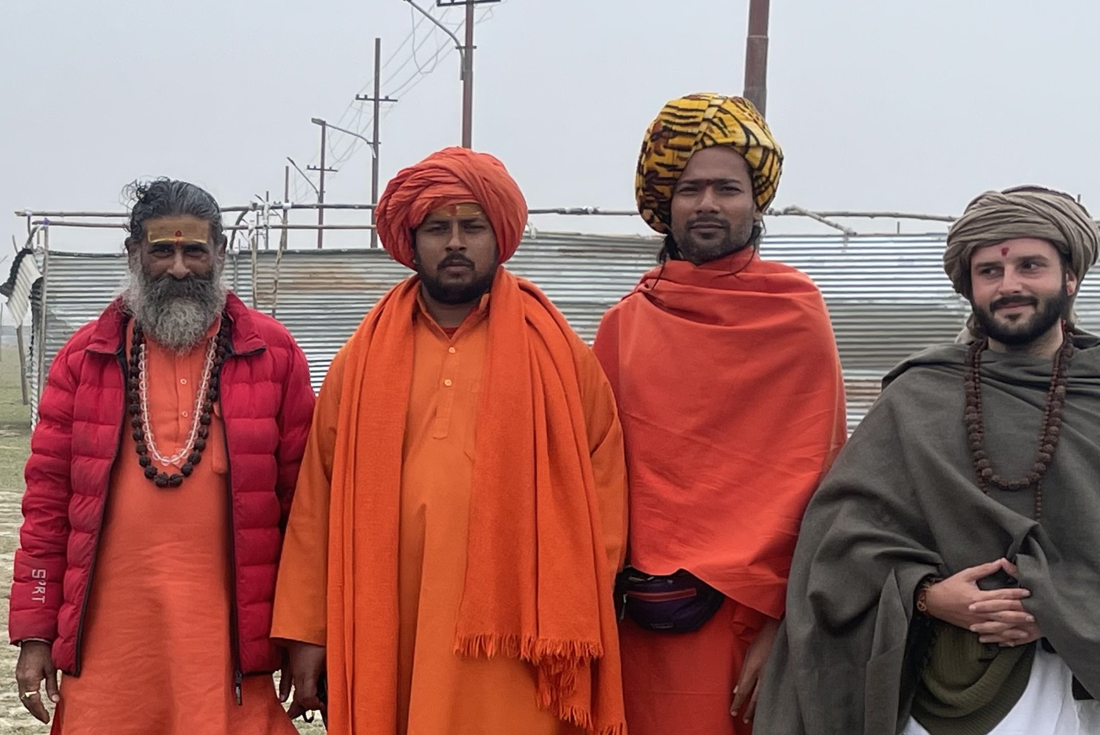 Four men dressed in traditional Indian spiritual and religious attire, standing outdoors in front of a metallic structure and utility poles.