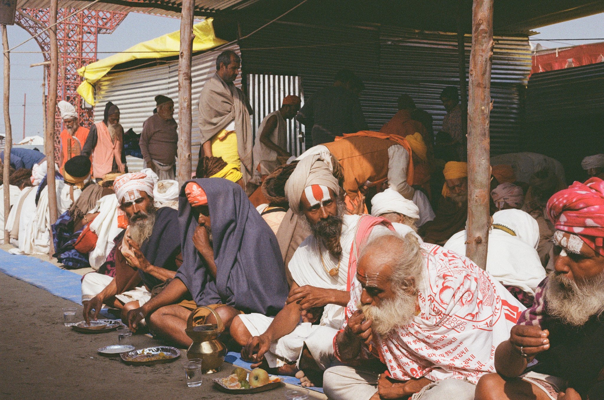 A group of men, many with beards and traditional Indian clothing, sitting on the ground in a line, possibly participating in a religious or cultural ceremony, with offerings and bowls of water and food items in front of them, under a tent structure.