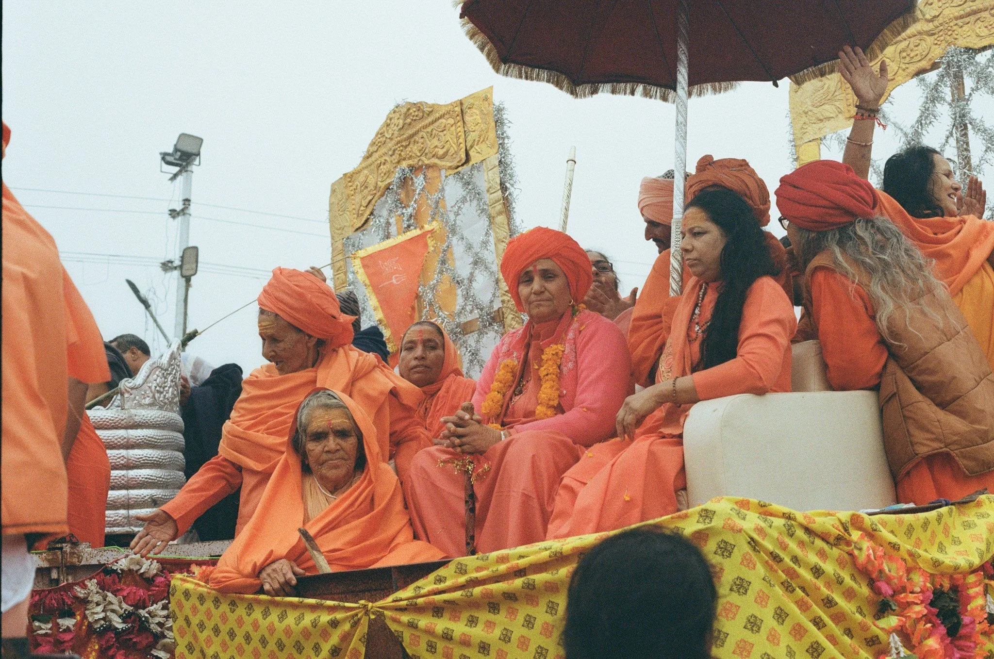 A group of Indian women dressed in orange and pink traditional attire sitting on a decorated float at a festival, with some wearing orange head coverings and others with garlands around their necks. The float is decorated with yellow cloth and flower