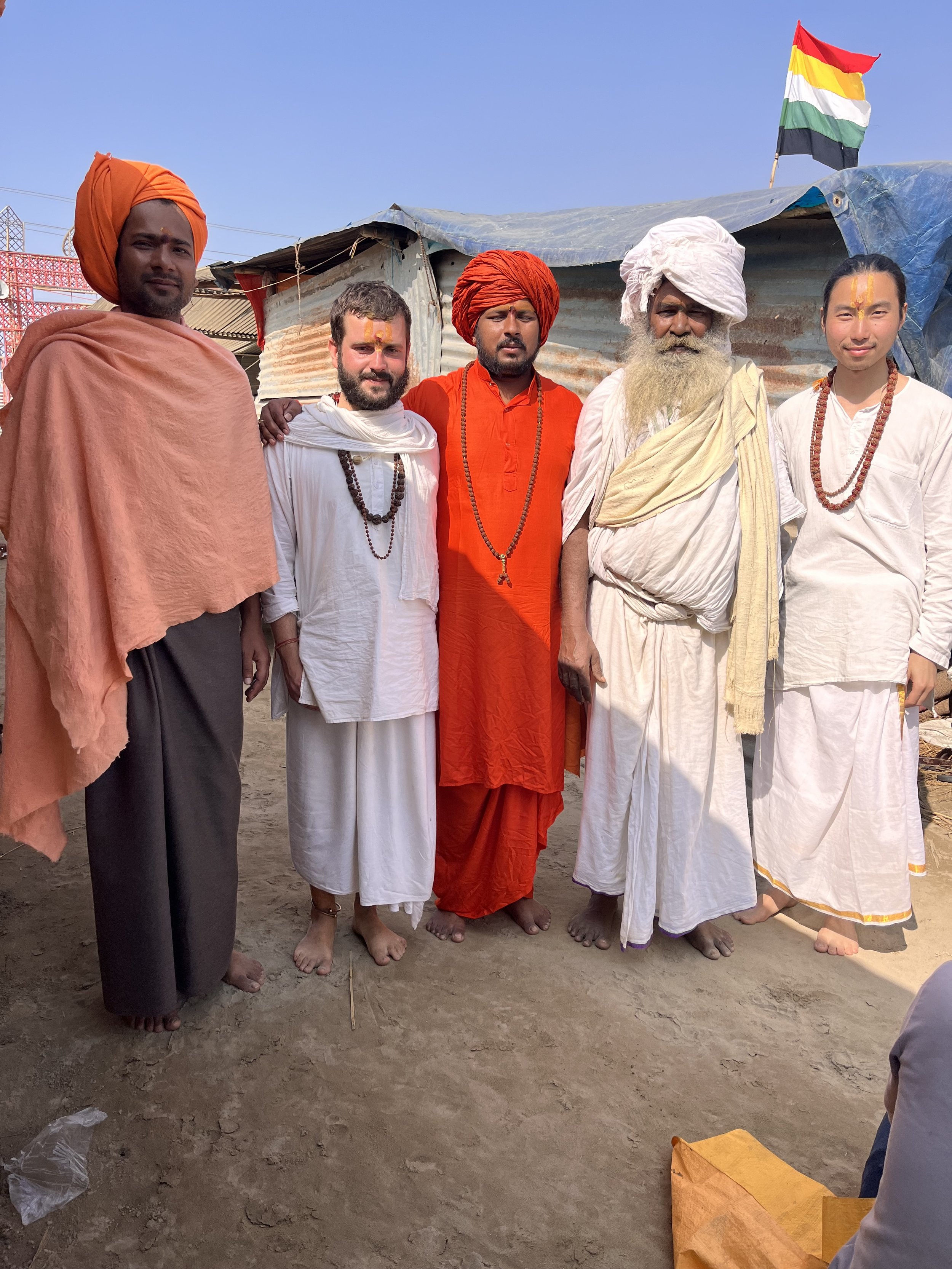 Group of six men dressed in traditional Indian attire, standing outdoors on dirt ground, with a temporary shelter and a flag in the background.