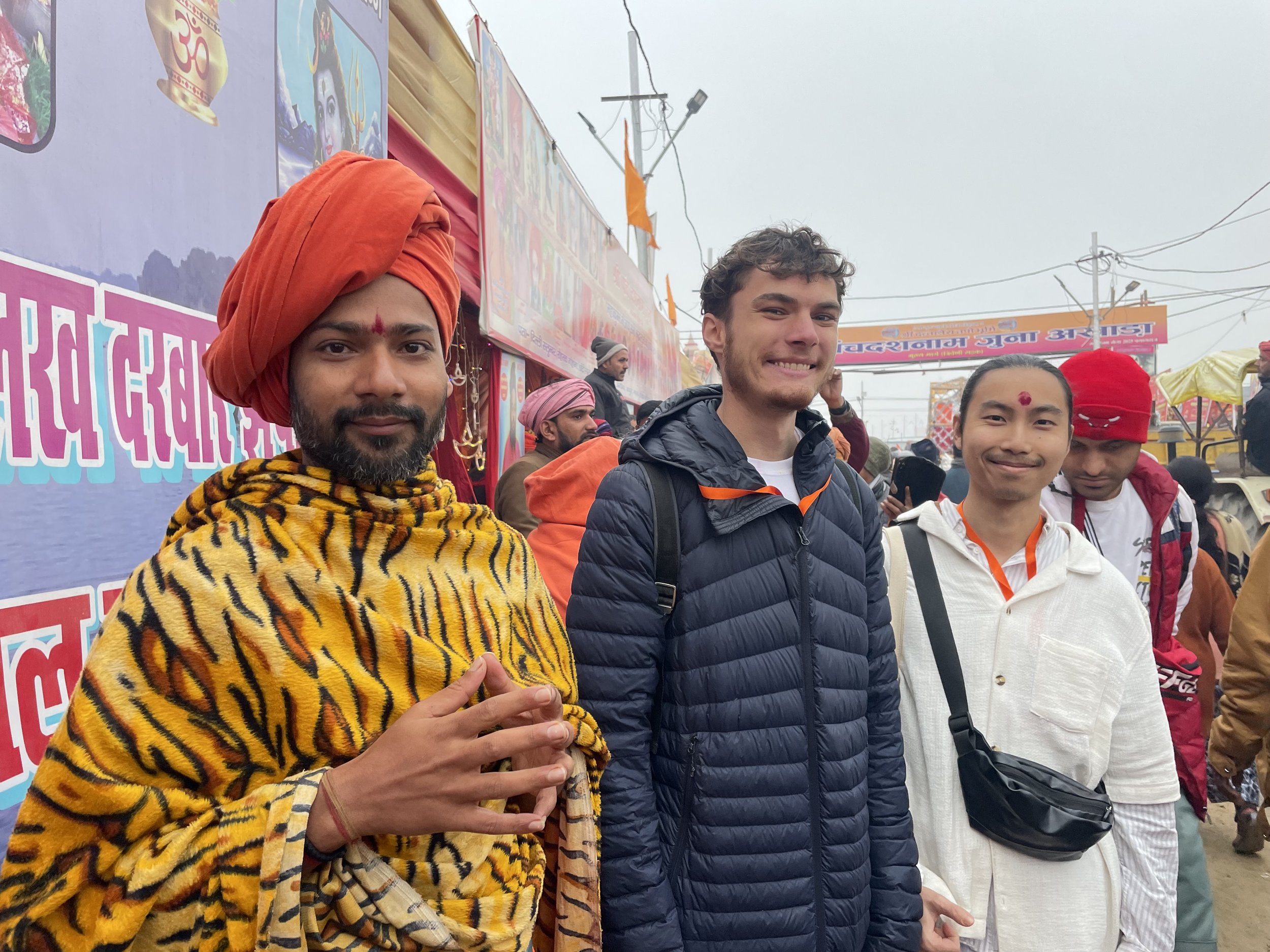 Four men smiling, standing outdoors at a festive event, with colorful banners and people in the background.