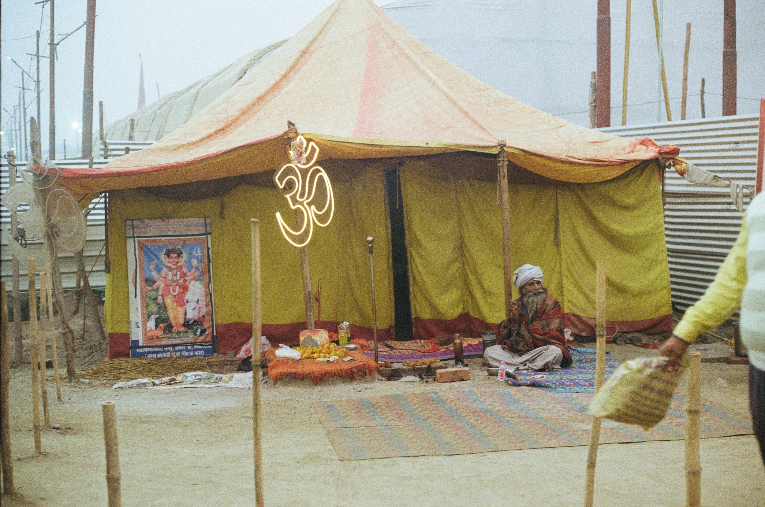 A yellow and orange tent set up outdoors with a man sitting on a colorful carpet, performing a ritual or prayer. There is a large poster of a Hindu deity in front of the tent, and various items for the ritual are placed on a small table. A glowing ne