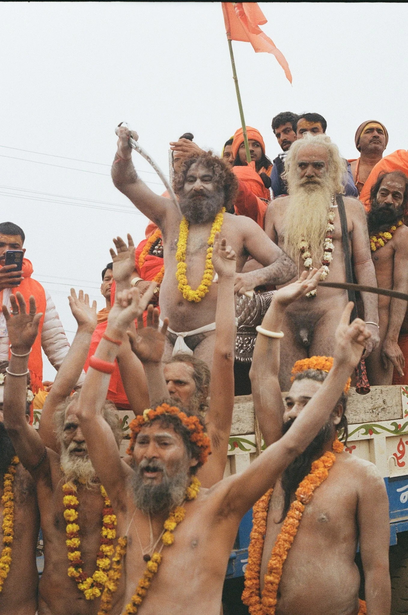 Group of men, many with long hair and beards, participating in a religious procession, some wearing marigold flower garlands. One man holds a yellow flag. The men are raising their hands and appear to be in a celebratory mood.