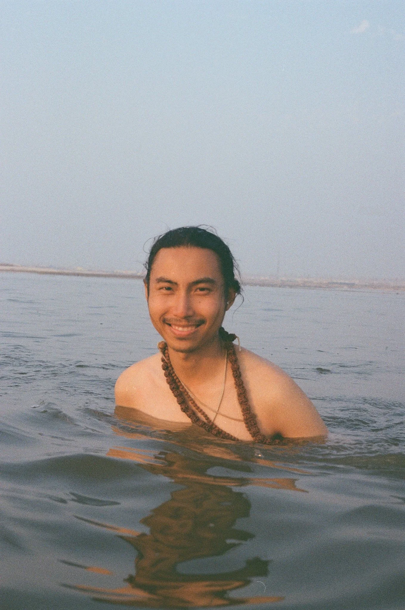 A smiling man with long hair tied back, wearing necklaces, swimming in the water with a body of water and a distant shoreline visible in the background during sunset or sunrise.