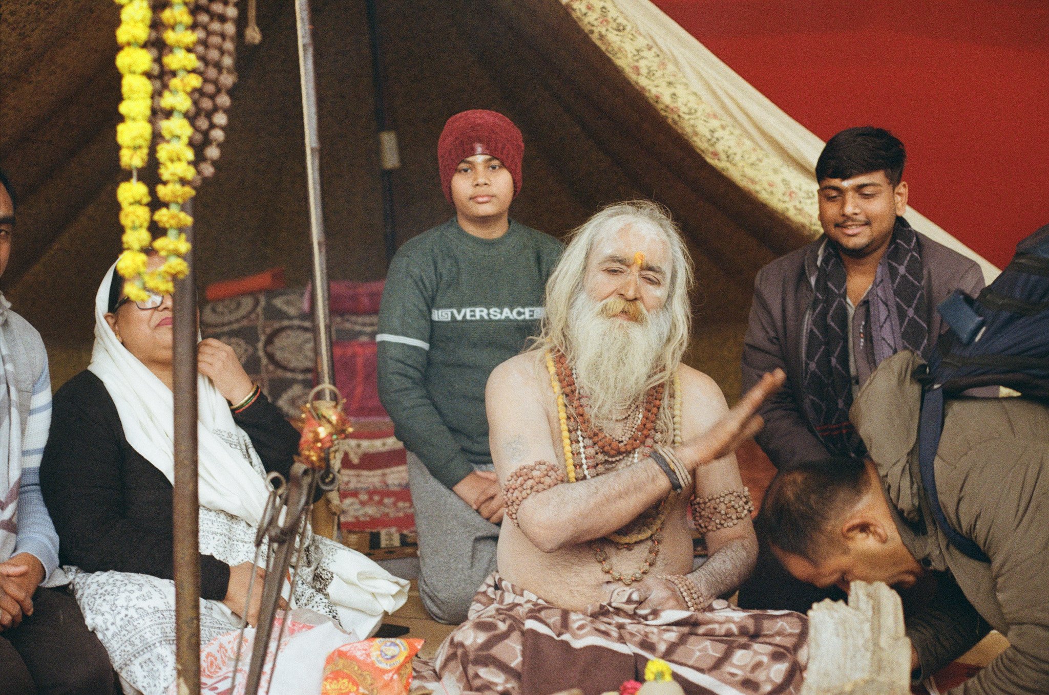 A group of people at a traditional spiritual ceremony inside a tent, with a revered elder who has long white hair and a beard, wearing multiple necklaces and traditional attire, performing a ritual or blessing.