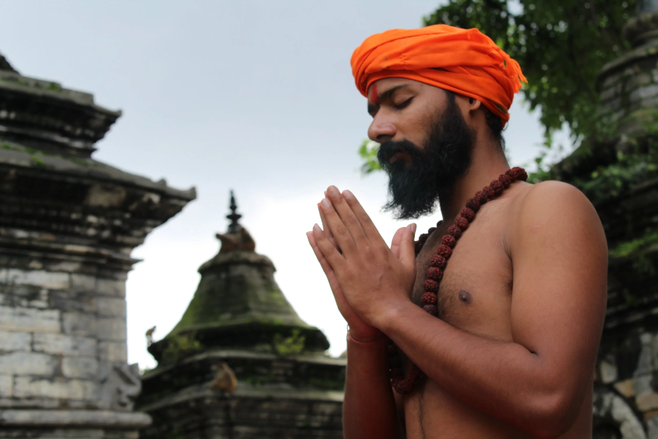 A man with a beard and orange turban, wearing a beaded necklace, stands shirtless with his hands pressed together in prayer, in front of ancient temple ruins with stone spires. yoga teacher, tantric practitioner, nepal