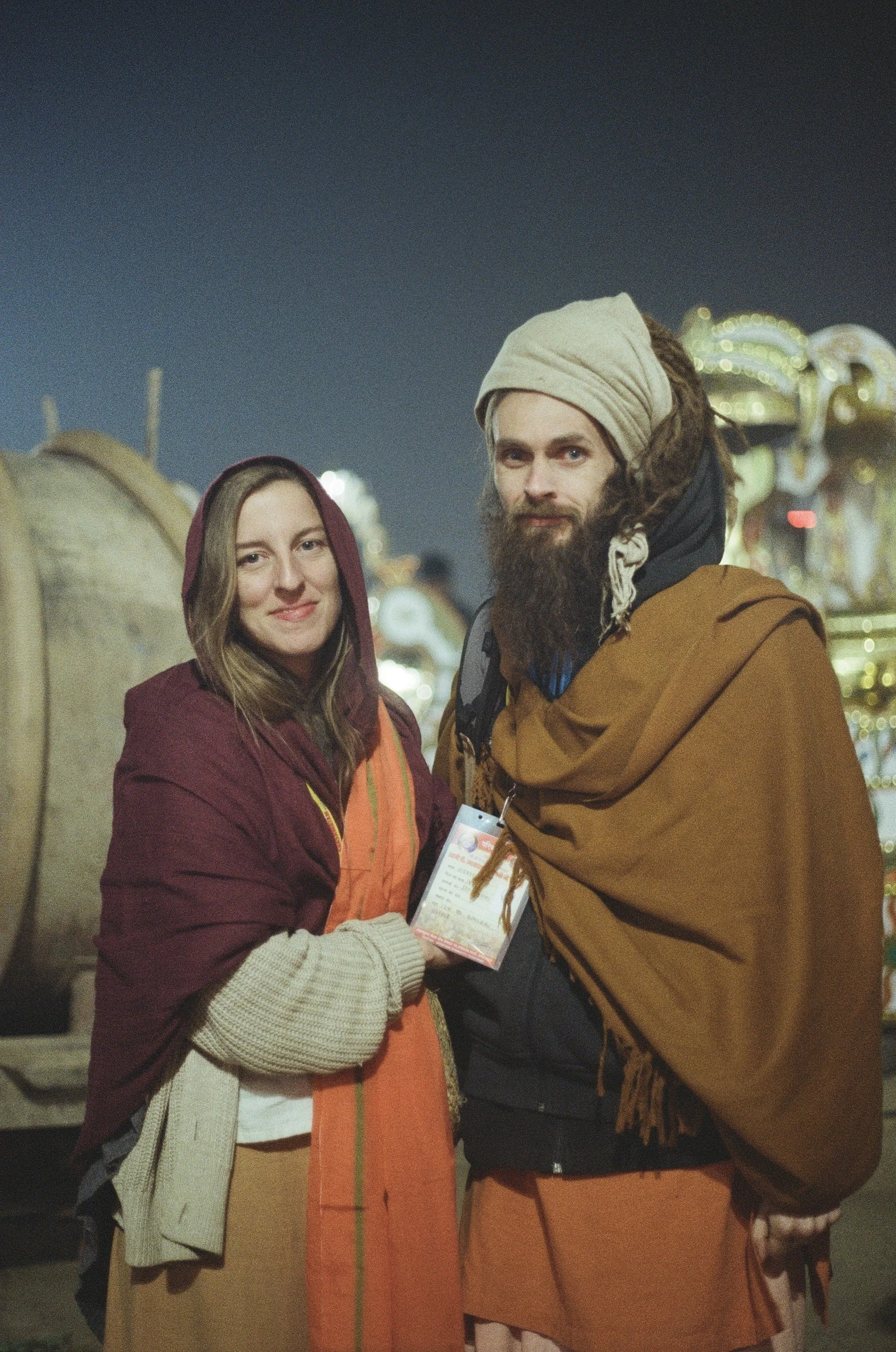 A woman and a man dressed in costume, standing together at night at an amusement park or carnival, with blurred lights and rides in the background.