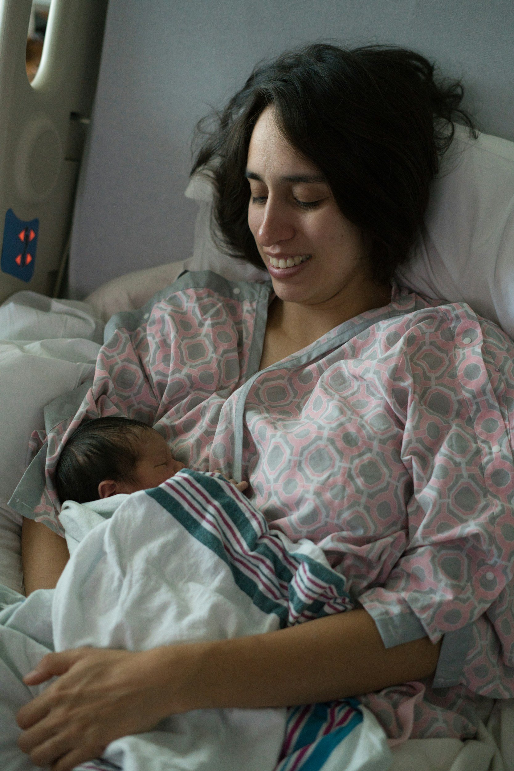 A woman in a hospital bed holding a newborn baby wrapped in a striped hospital blanket, looking at the baby with a smile.