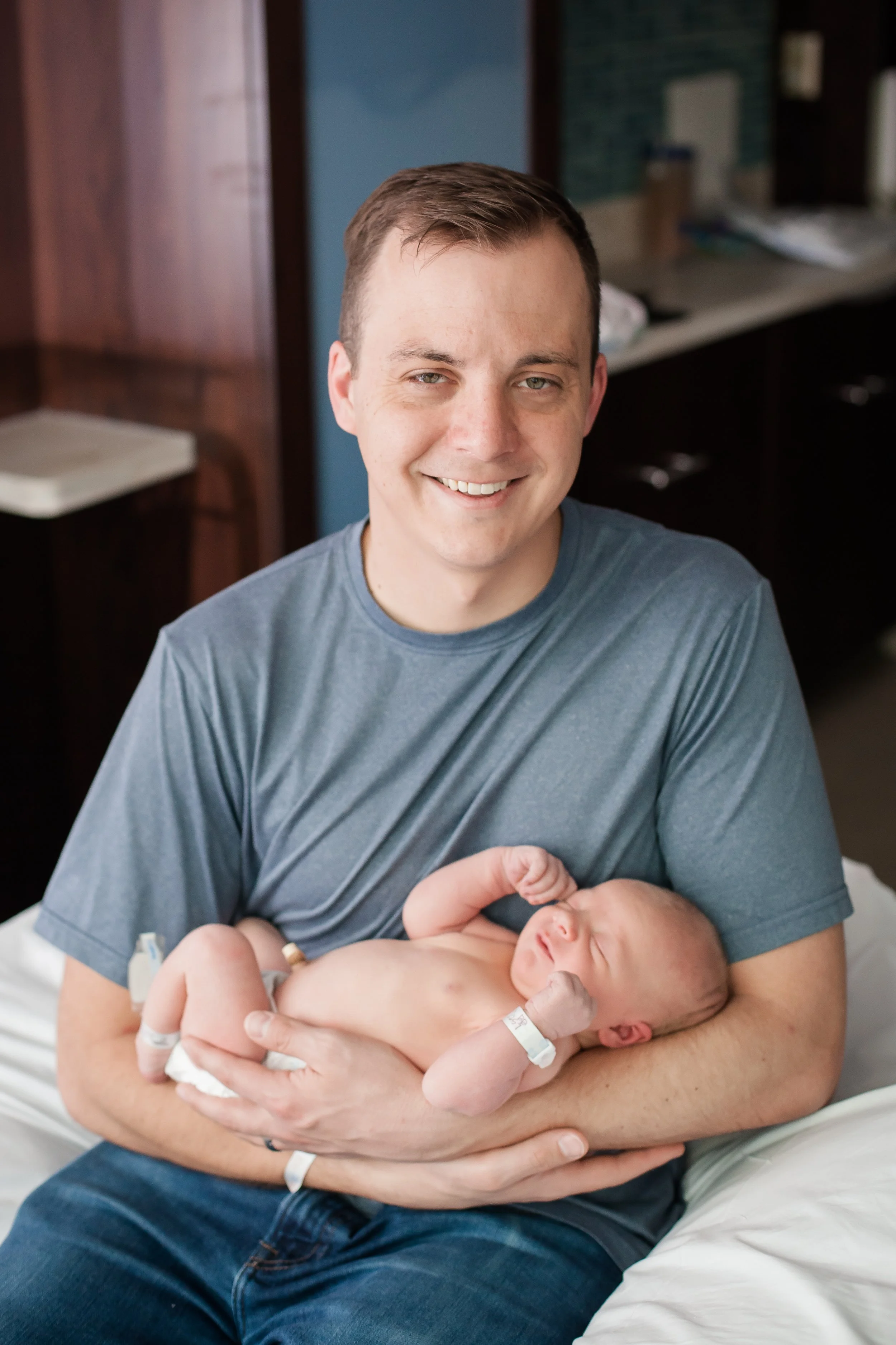 A man in a gray T-shirt holding a newborn baby in a hospital room.