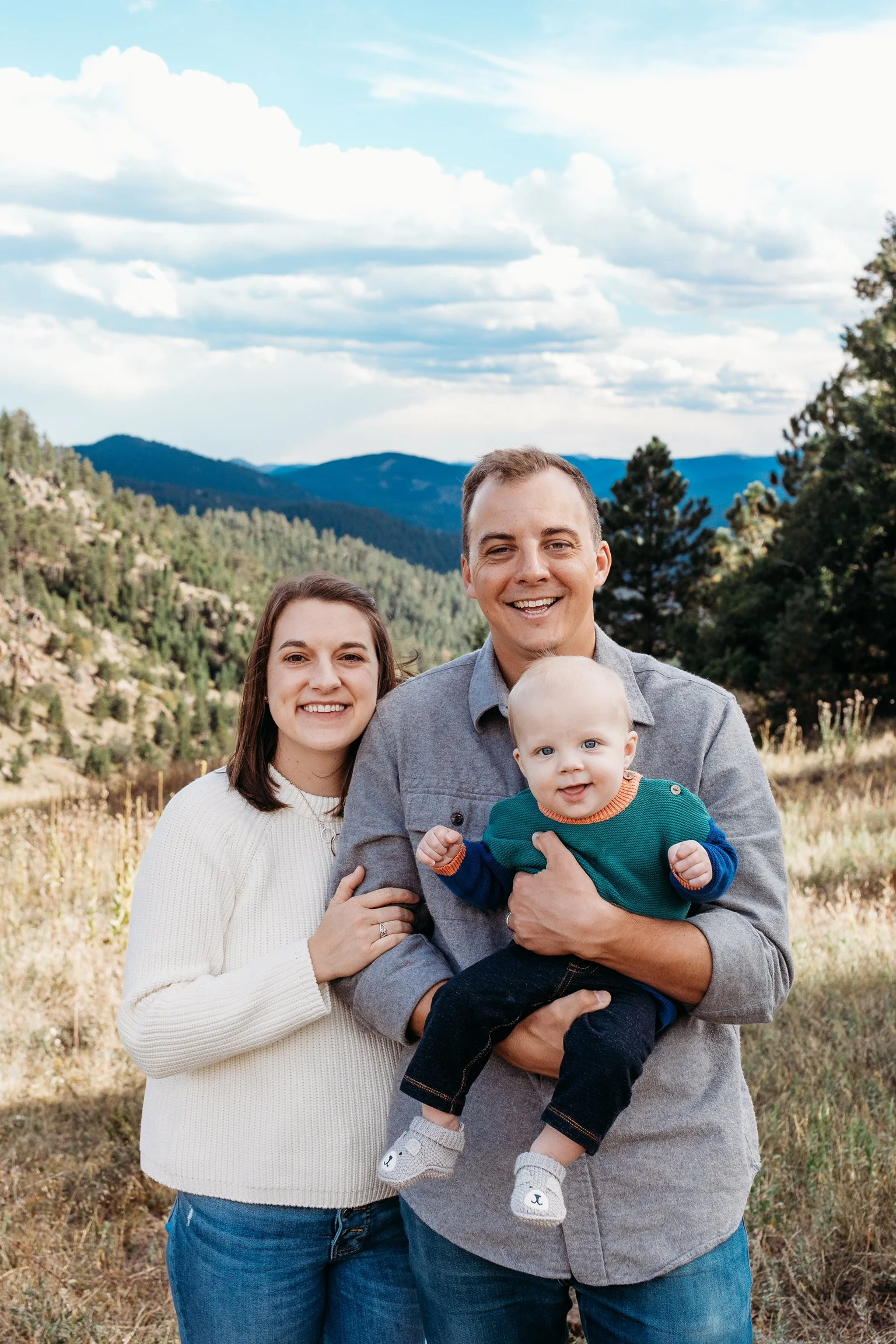 A happy family of three standing outdoors in a mountainous area with trees and a partly cloudy sky. The man is holding a smiling baby boy, and the woman stands beside them, smiling at the camera.