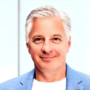 Michael Kaune mit weißen Haaren, trägt eine blaue Jacke und lächelt. Im Hintergrund ist ein hell erleuchtetes Fenster.