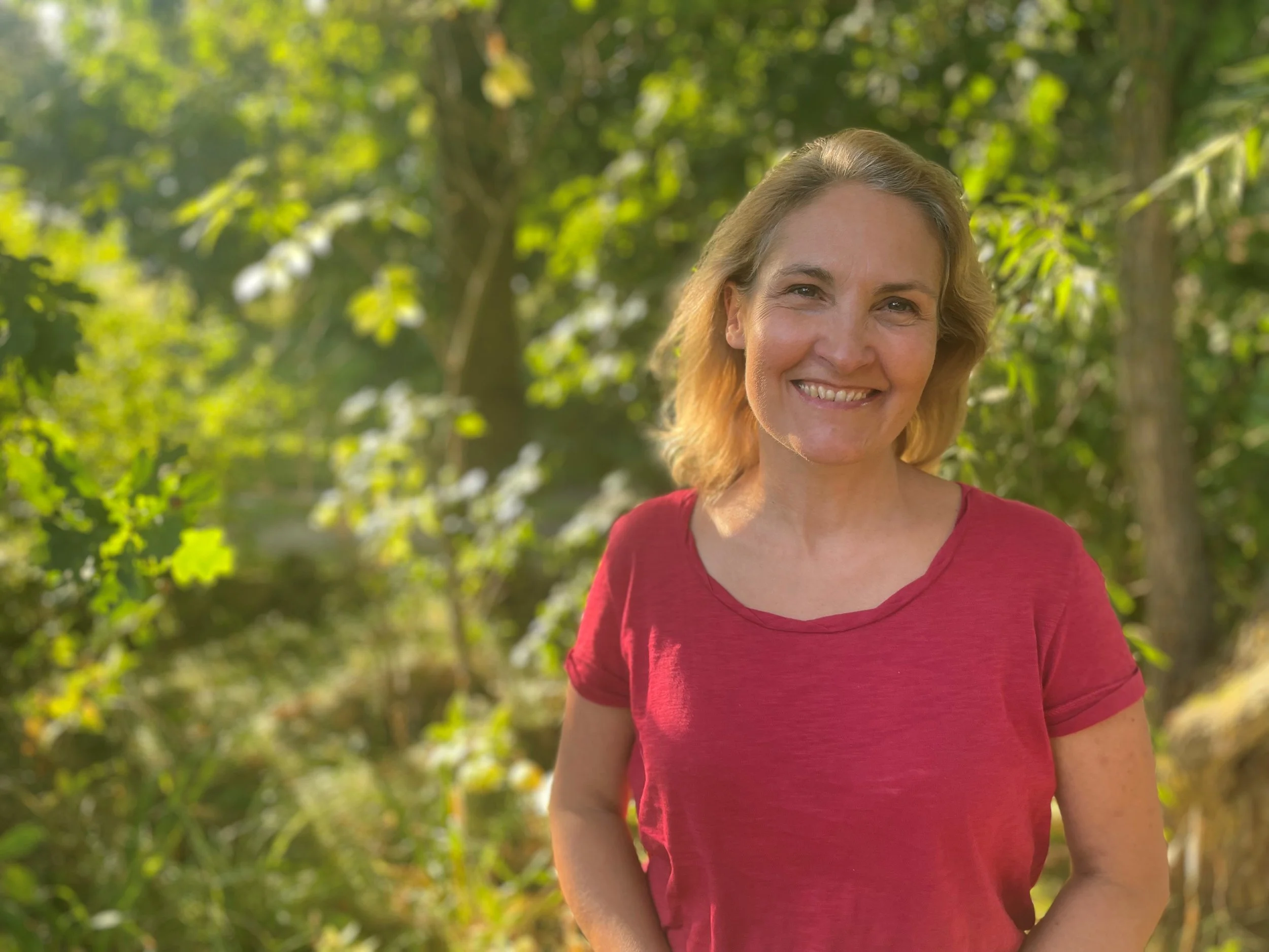 Marion Leo in einem roten T-Shirt steht in einem grünen Wald, Sonnenlicht fällt durch die Bäume.