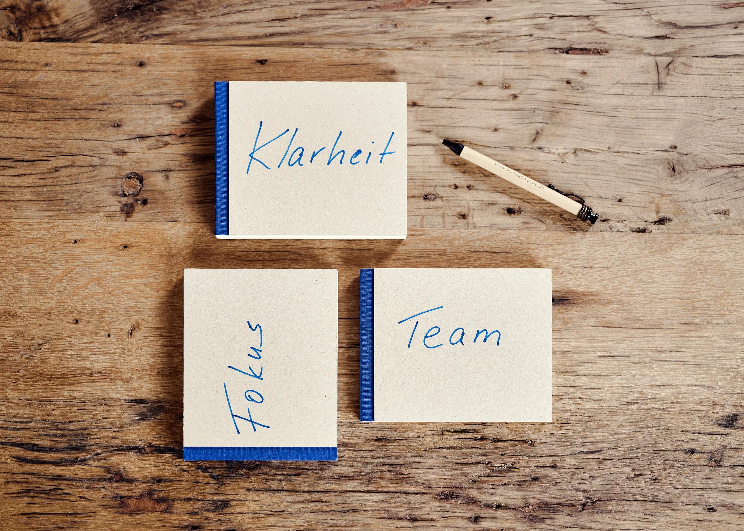 Drei kleine Notizbücher mit blauer Schrift auf Holzoberfläche, eines mit 'Klarheit', eines mit 'Team', eines mit 'Fokus', und ein schwarzer Kugelschreiber.