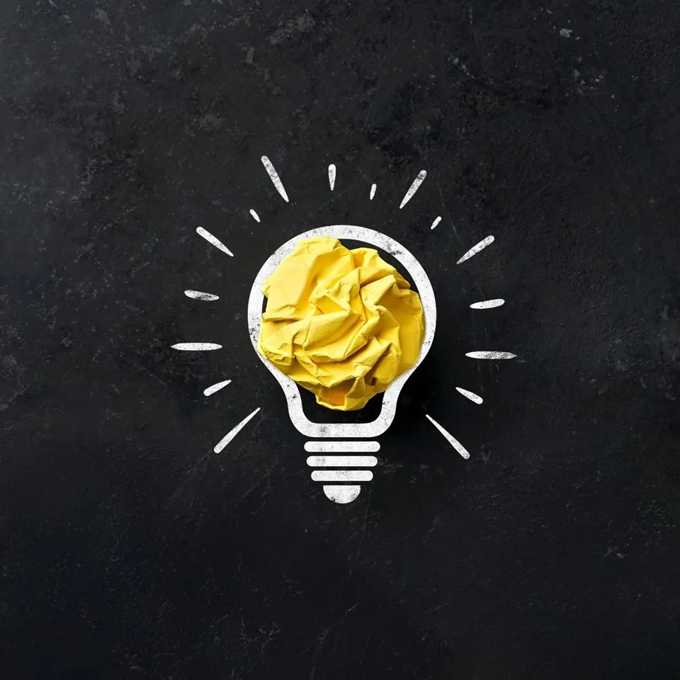 Eine Zeichnung einer Glühbirne auf einer schwarzen Oberfläche, mit zerknülltem gelbem Papier in der Mitte der Glühbirne.