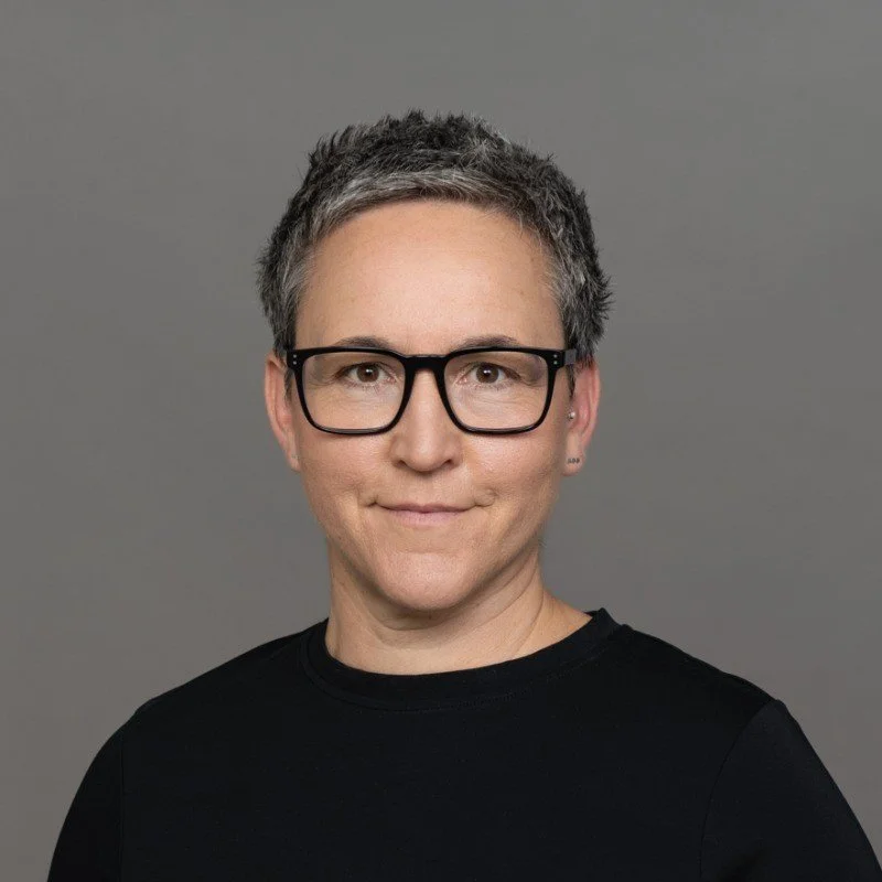 Julia Fernández-Pola mit kurzen, grau melierten Haaren, schwarzen Brillen und schwarzem Oberteil vor grauem Hintergrund.