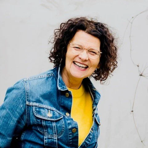 Elke Marie Ekert lächelnd, mit Brille und lockigem Haar, trägt gelbes Sweatshirt und eine Jeansjacke, vor weißer Wand.