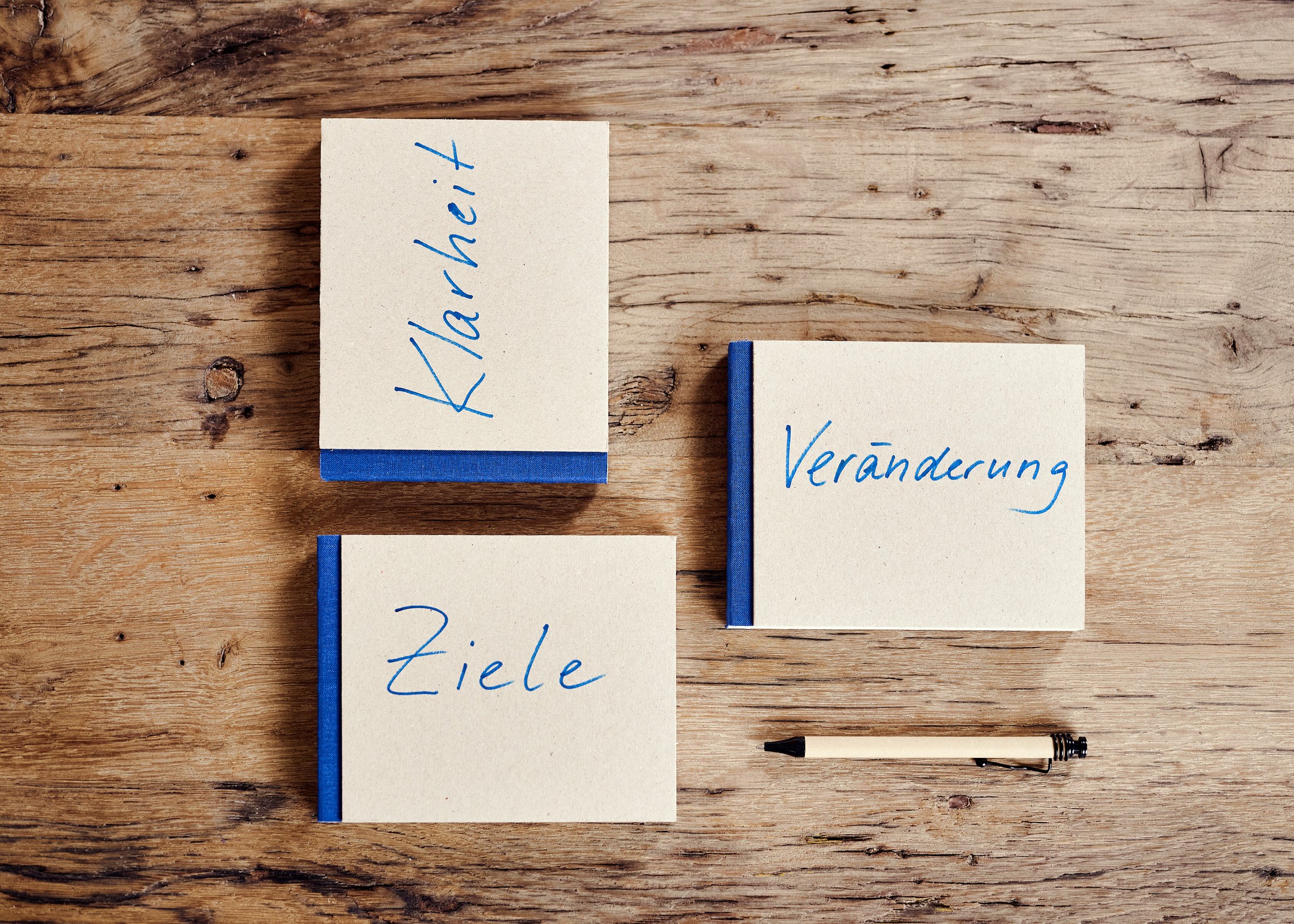 Drei Notizbücher mit blauer Bindung und handgeschriebenen Wörtern in blauer Schrift auf einem Holztisch, daneben ein Kuli. Die Wörter sind 'Klarexh', 'Ziele' und 'Veränderung'.