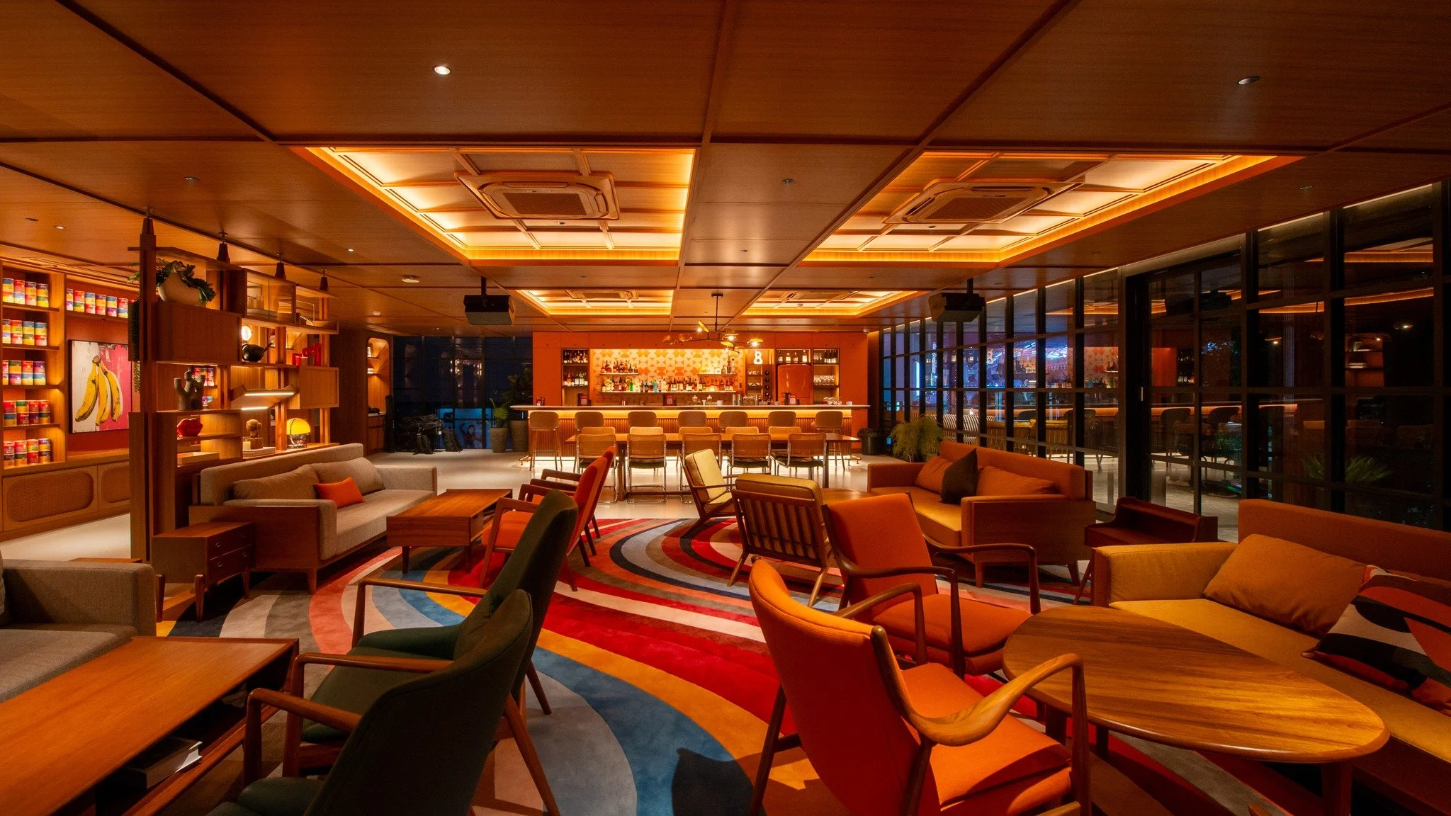 Warmly lit bar and lounge area with colorful furniture, wooden tables, and a patterned carpet, featuring a well-stocked bar in the background.