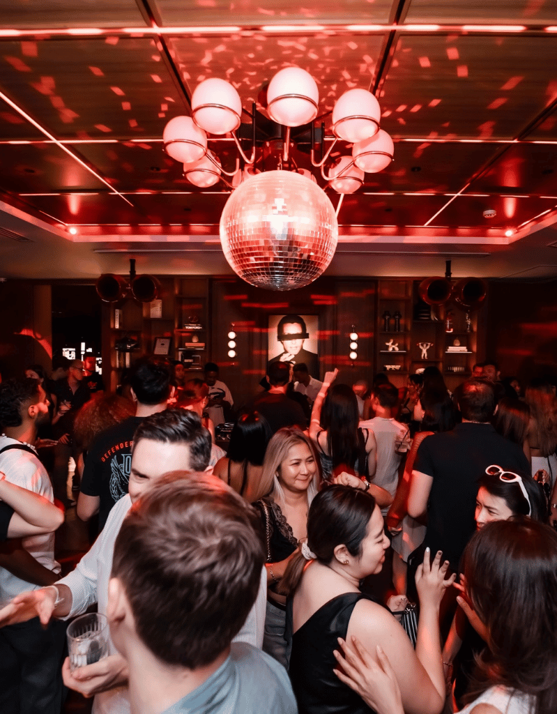 People dancing and socializing in a club with red lighting and a disco ball hanging from the ceiling.
