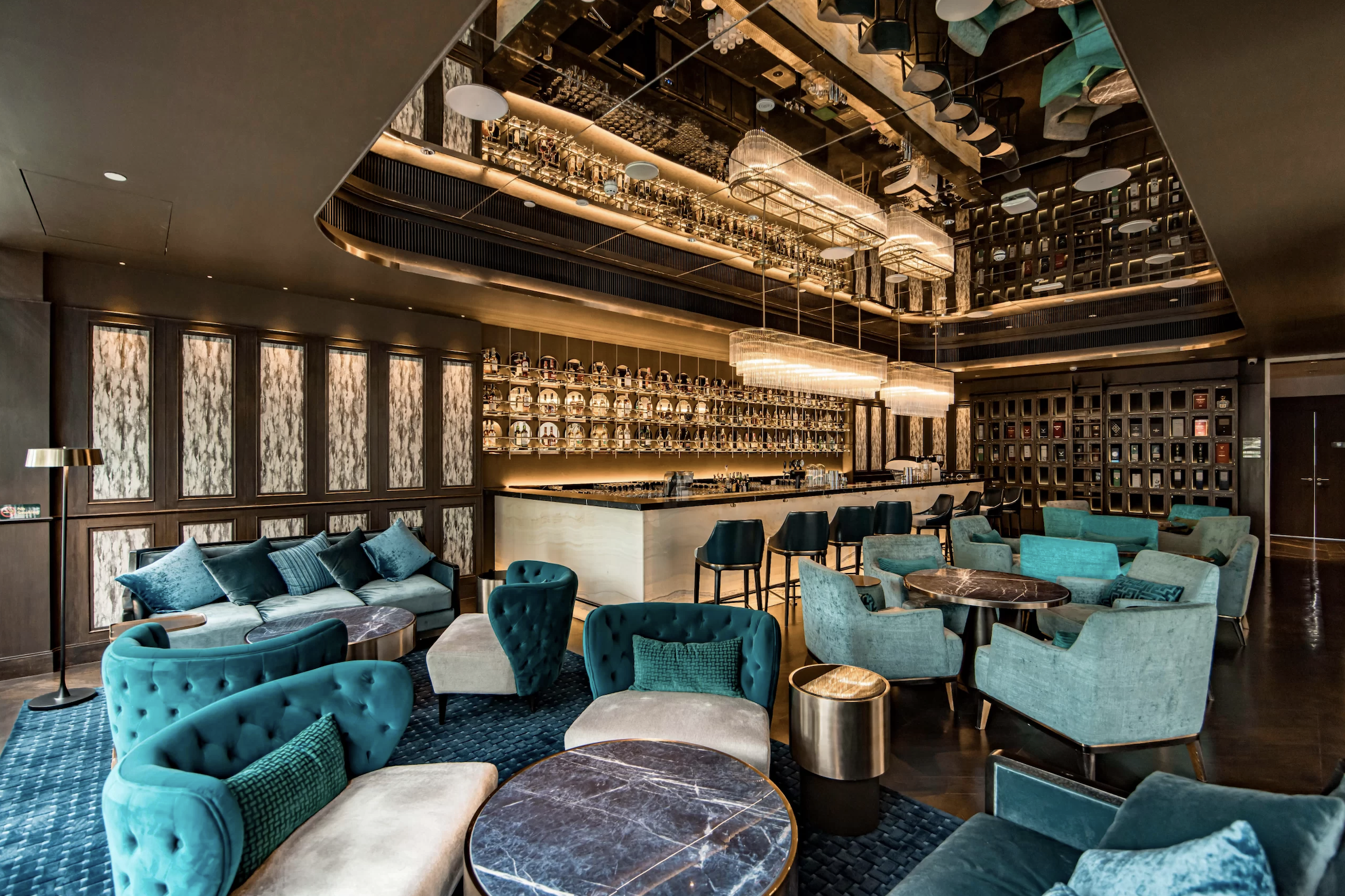 Interior view of a luxurious lounge bar with modern decor, teal and beige upholstered seating, marble-topped tables, a well-stocked bar with bottles displayed on shelves, and warm ambient lighting.