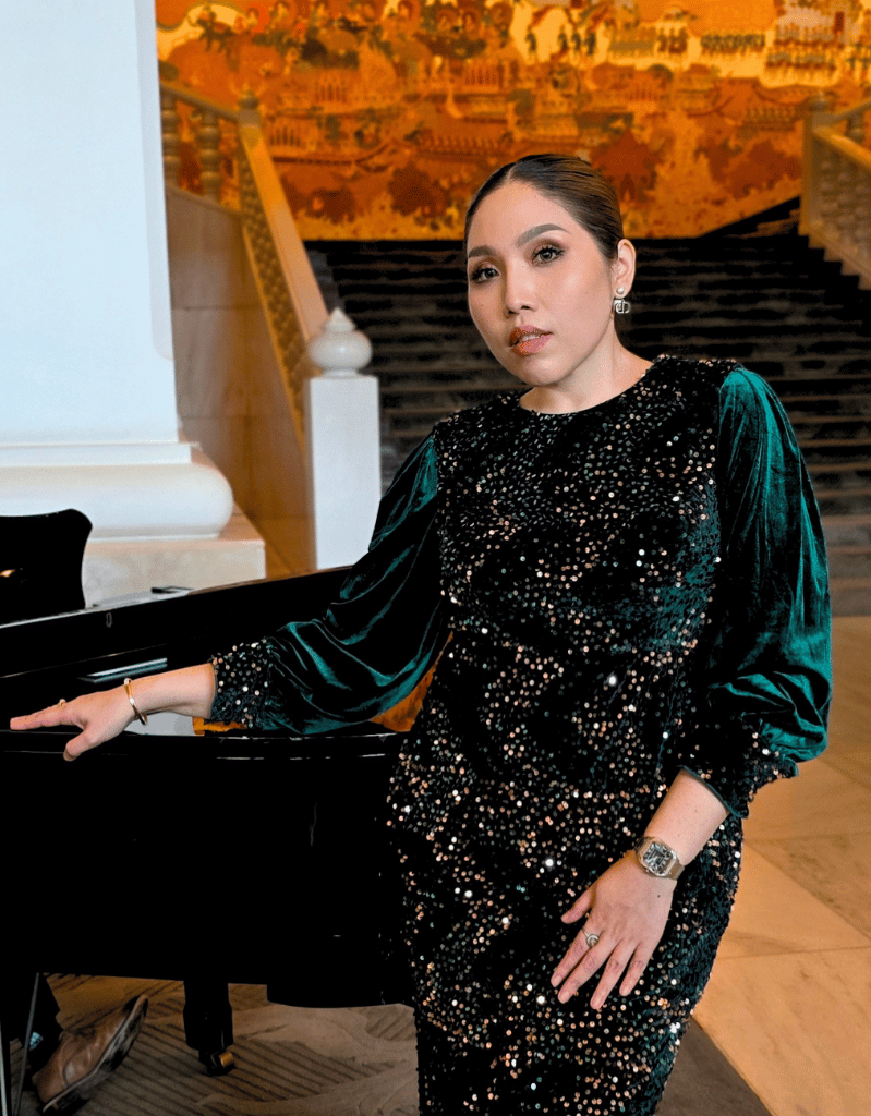 A woman wearing a black dress with sparkling embellishments, sitting at a black grand piano in a decorated interior space with a gold and orange mural in the background.
