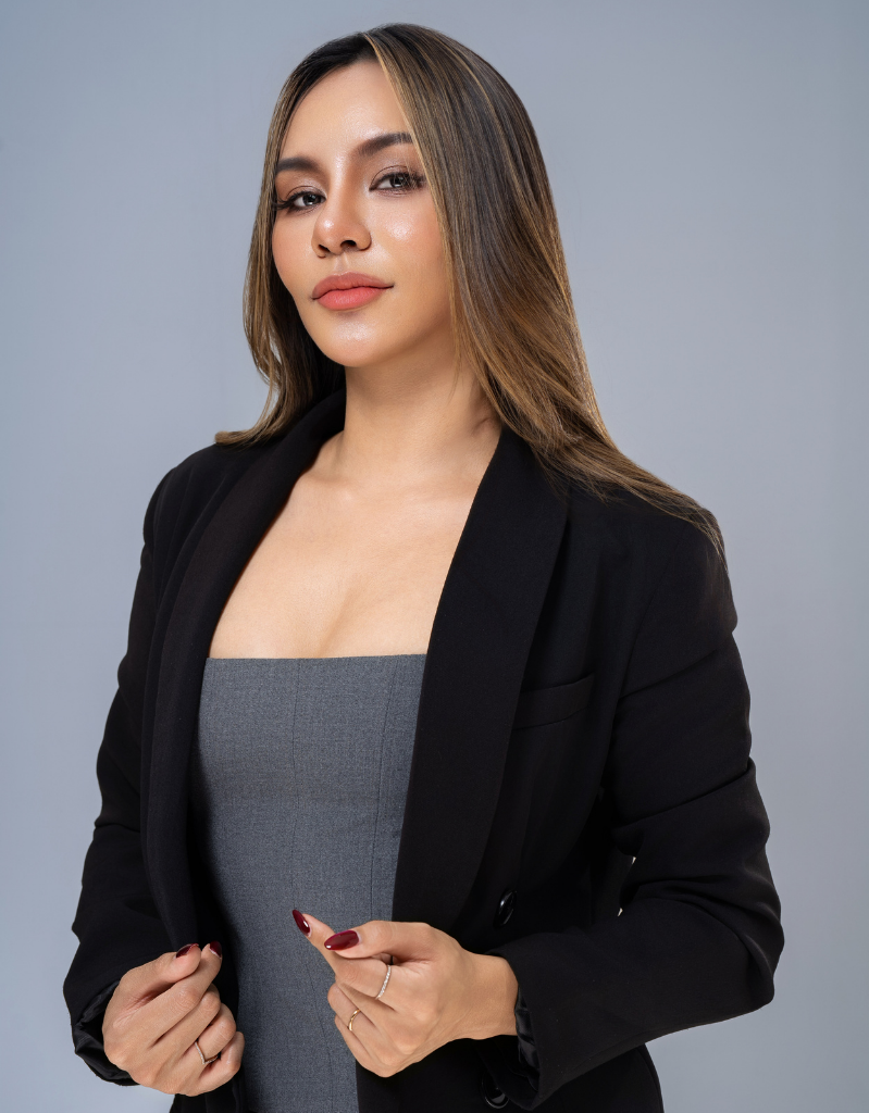 A woman with straight brown hair, wearing a gray top and black blazer, posing confidently with a slight smile against a light gray background.