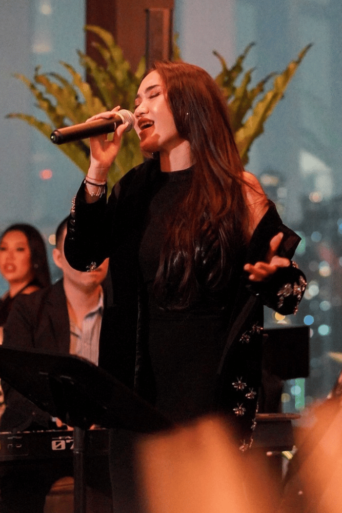 A woman singing into a microphone at an indoor event, with a keyboard and a woman in the background. She has long dark hair and is wearing black clothing.