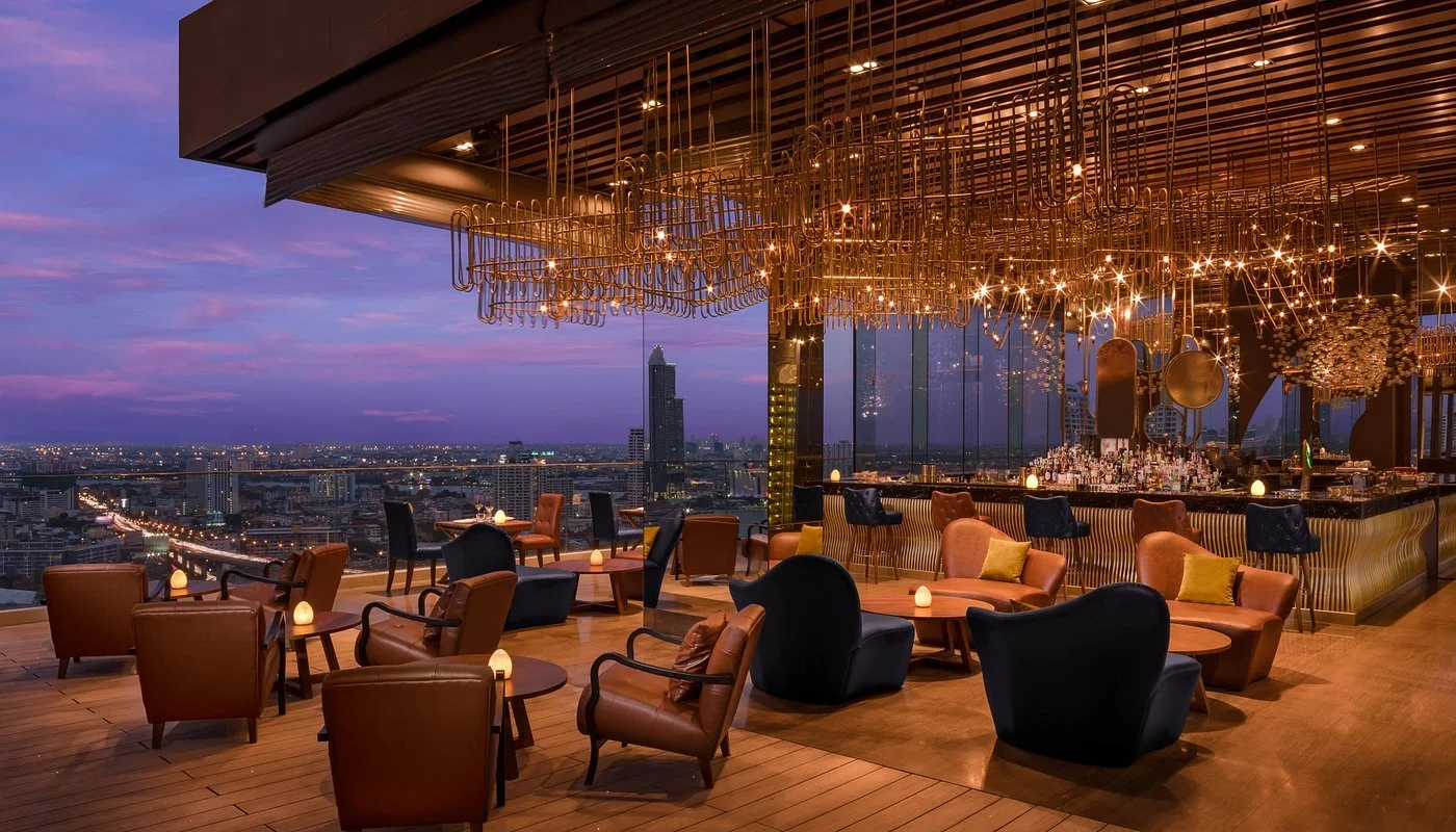 Upscale rooftop restaurant and bar overlooking a city skyline at sunset, with modern chandelier lighting, leather and fabric chairs, small tables with candles, and a bar area with bottles and glasses.