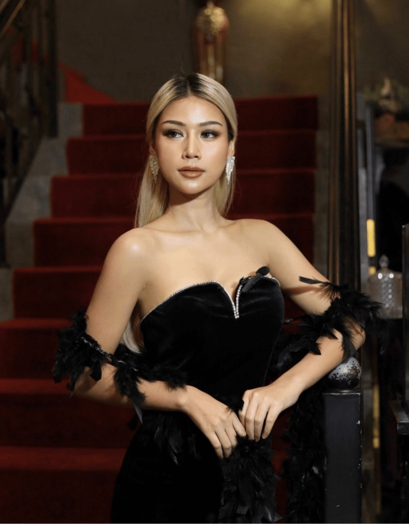 A woman with long blonde hair and earrings wearing a black strapless dress with feather details, standing near a staircase with red carpet.