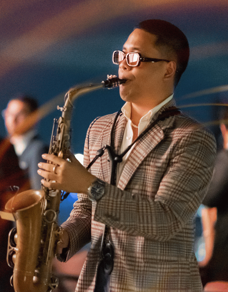 A man playing the saxophone at an indoor event, wearing sunglasses, a checkered blazer, and a watch.