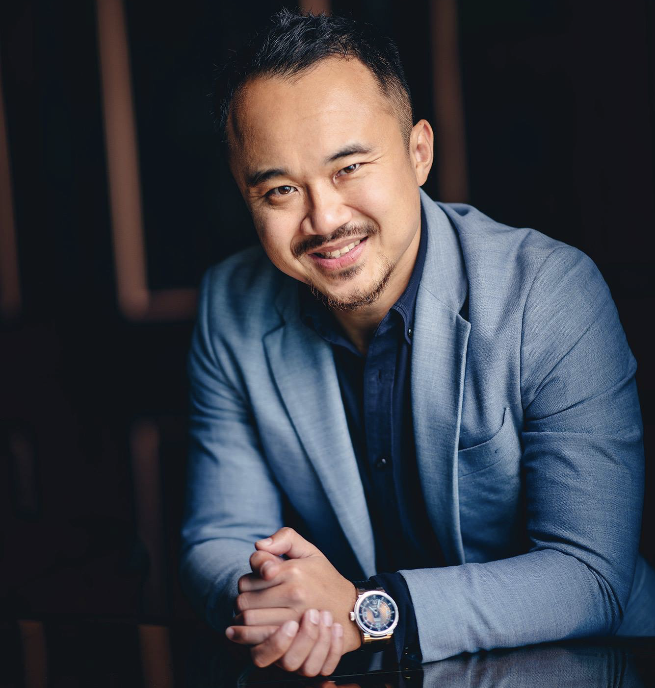 A man with short dark hair, facial hair, and a friendly smile, wearing a light blue blazer over a dark shirt, sitting at a table with his hands clasped, wearing a watch on his left wrist, in a dimly lit setting.