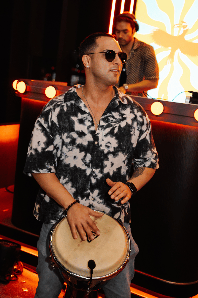 A man wearing sunglasses, a black and white floral shirt, and a watch playing a hand drum at a nightclub with colorful lighting and a DJ in the background.