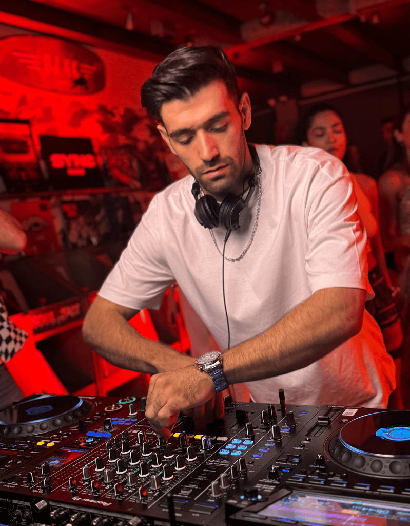 A male DJ wearing a white T-shirt and headphones around his neck, adjusting controls on a DJ mixer at a club with red lighting.
