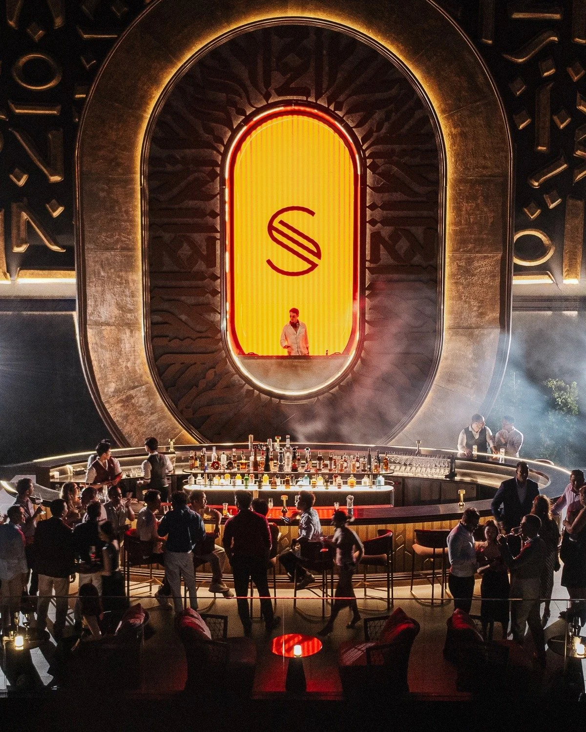 A crowded bar or nightclub with a large stage and a DJ or performer behind a glowing orange and yellow logo with a stylized 'S' symbol. People are socializing and enjoying drinks.