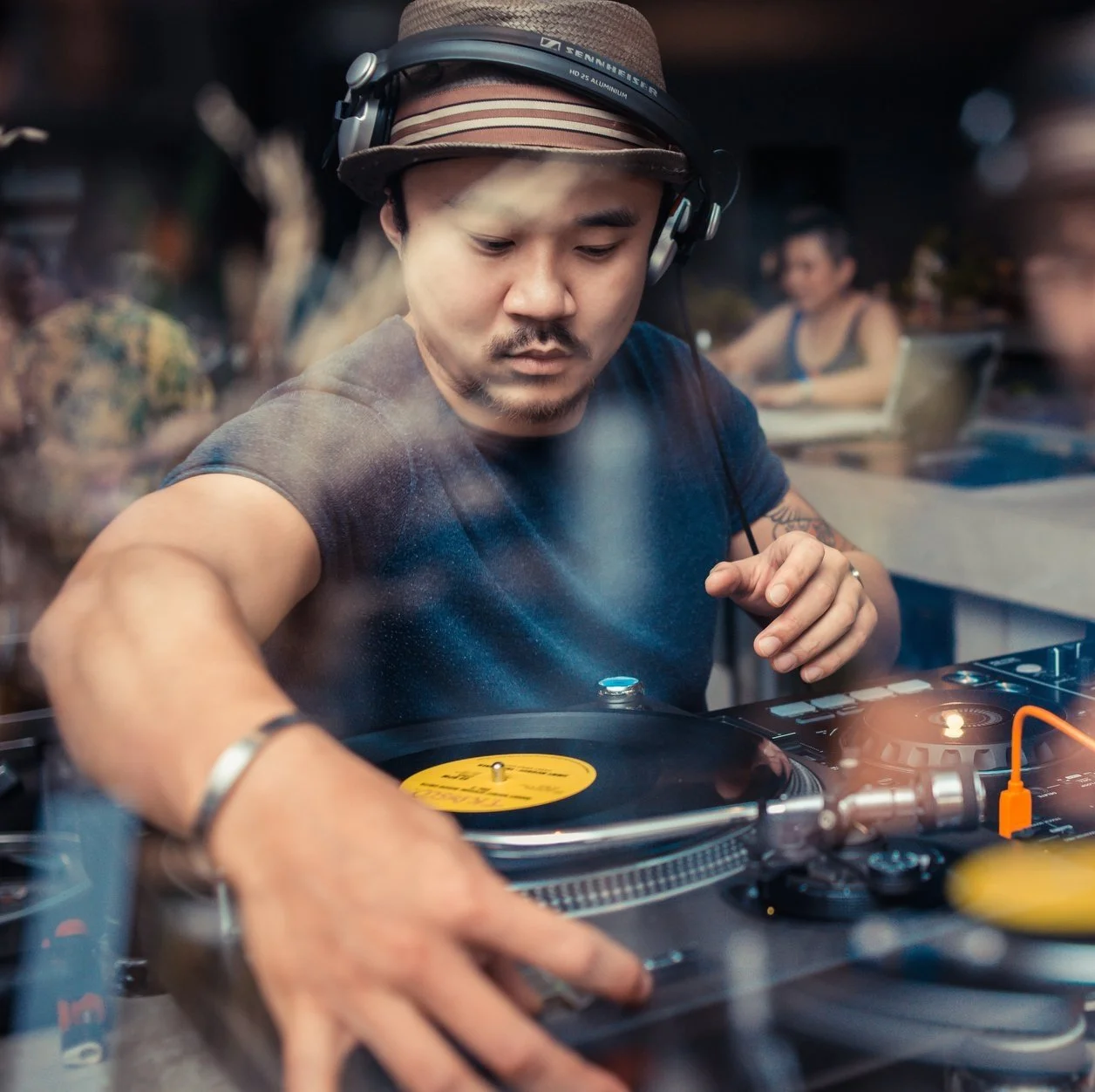 A DJ wearing a hat and headphones, focused on mixing music at a booth with turntables, in a lively setting with people in the background.