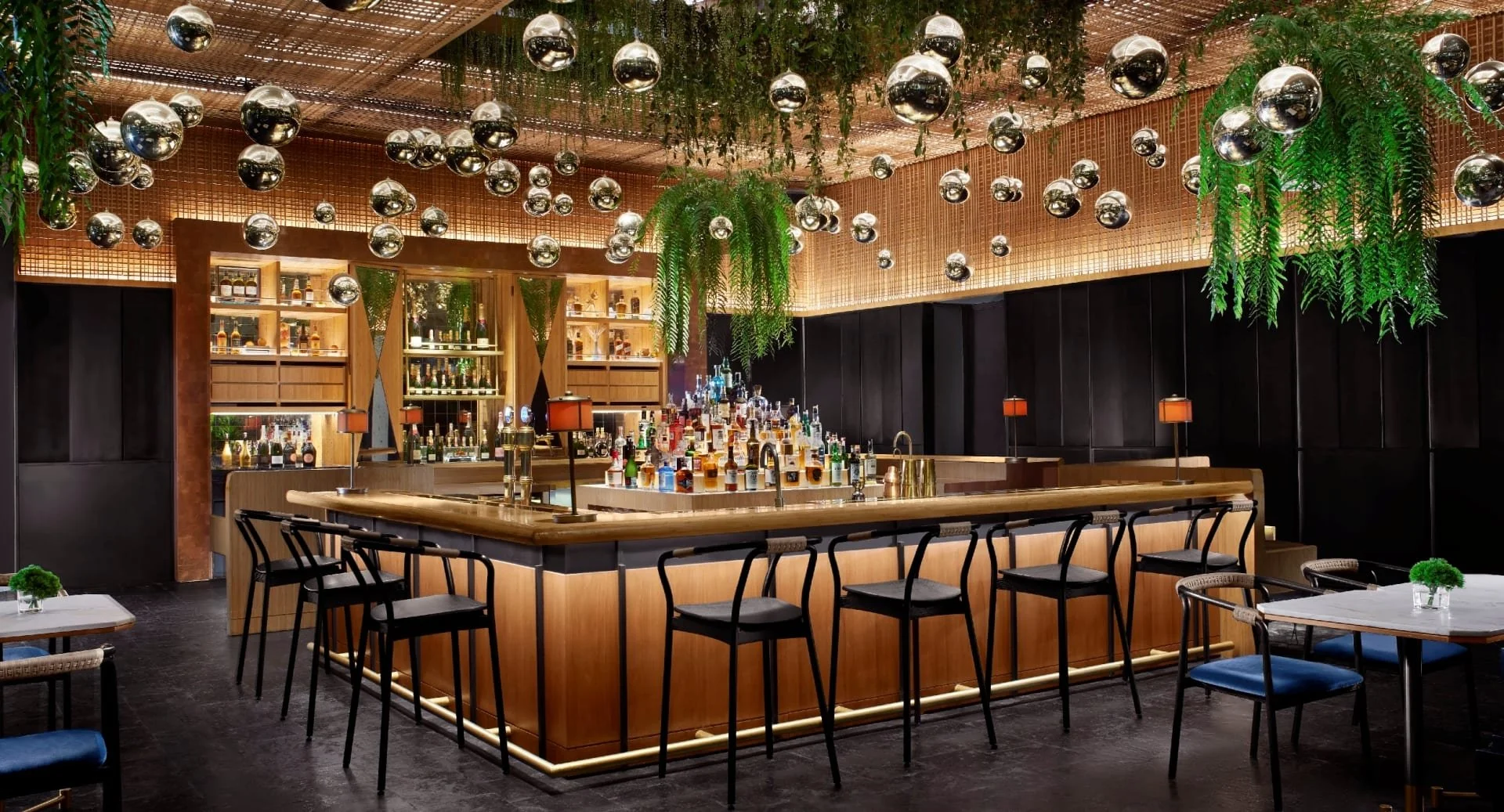 Interior of a stylish bar with a wooden counter, shelves filled with bottles, hanging greenery, and a ceiling decorated with reflective silver spheres and hanging plants