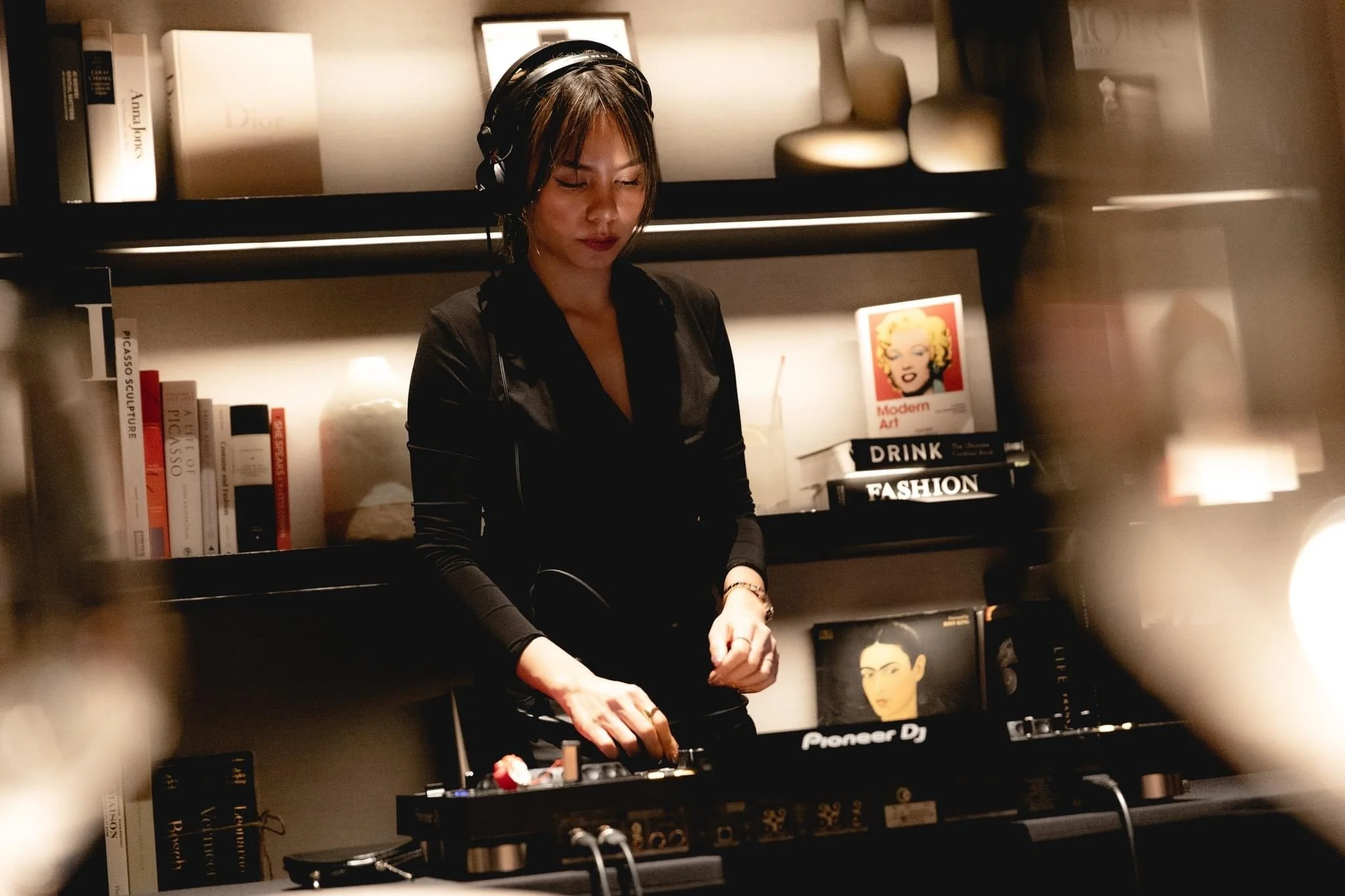 A female DJ wearing headphones is playing music behind a DJ mixer in a cozy, well-lit room with shelves of books and art décor.