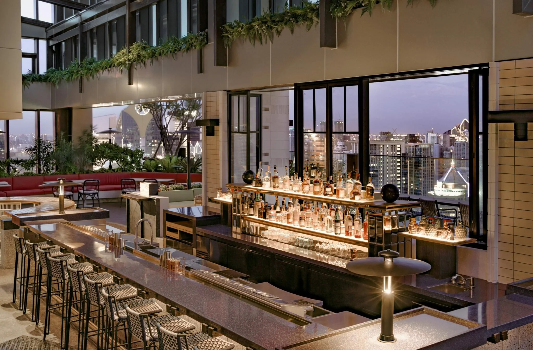 Upscale rooftop bar with a city skyline view, featuring a well-stocked bar counter with liquor bottles, a long seating area with patterned stools, and modern decor with plants and large windows.