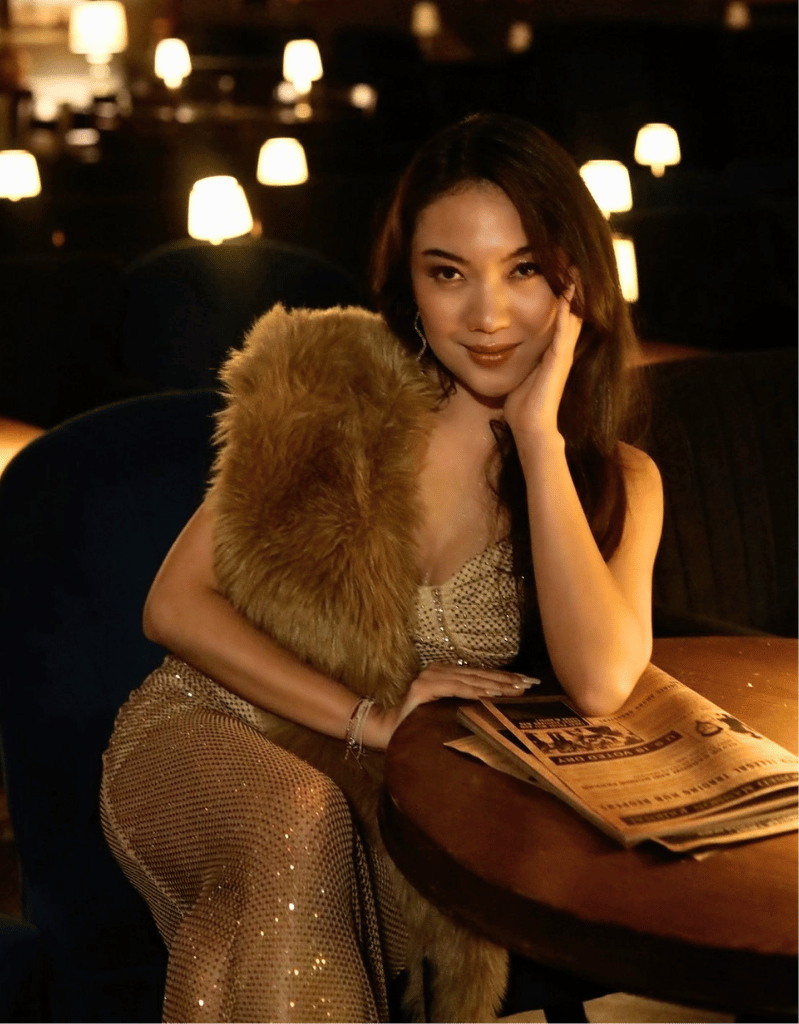 A woman sitting at a table in a dimly lit restaurant or lounge, with small lamps in the background. She is wearing a sparkly dress and a fur stole, posing with one hand on her face and a subtle smile.