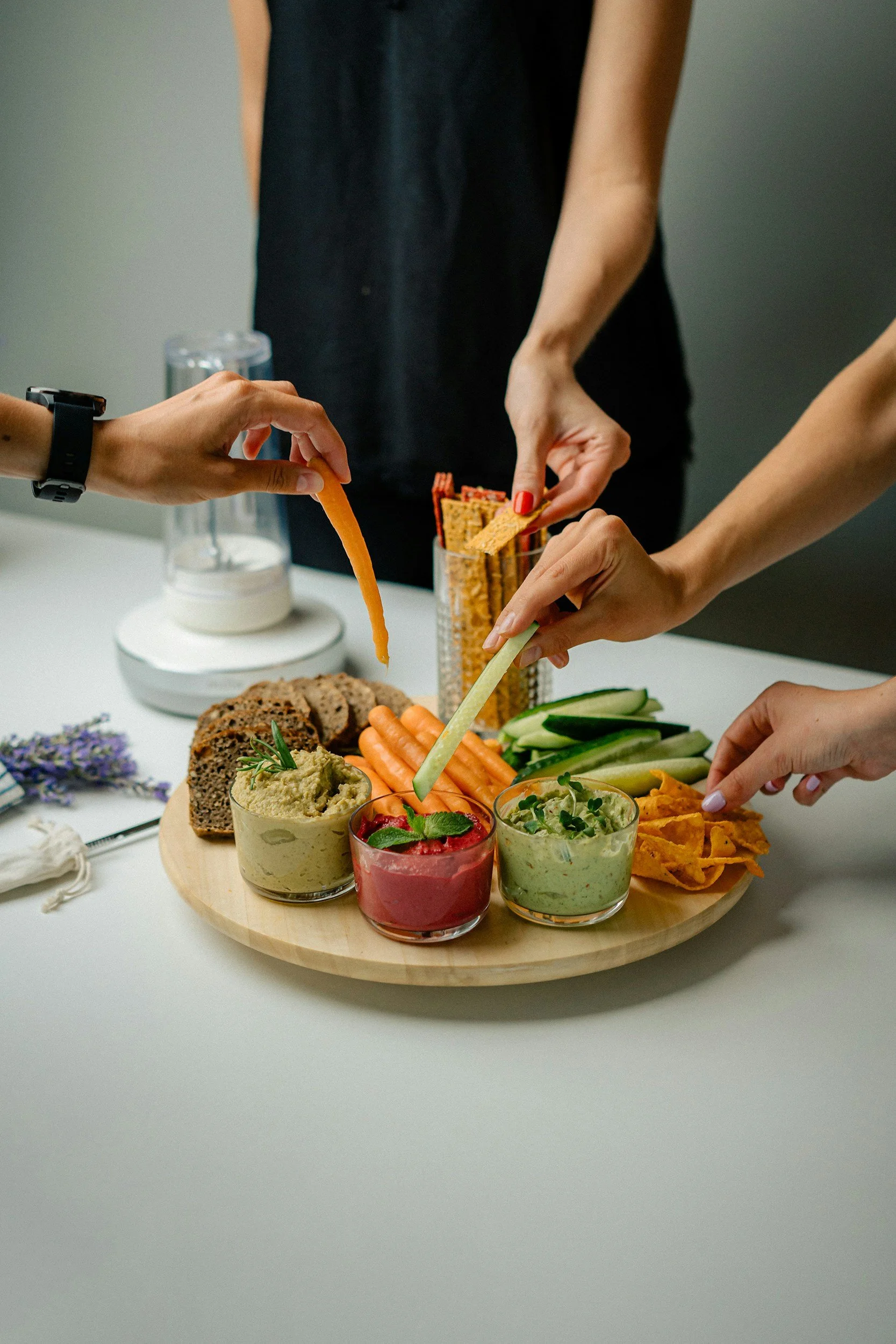 People place vegetables and dip on a round wooden platter, with hummus, guacamole, salsa, carrots, cucumbers, and chips.