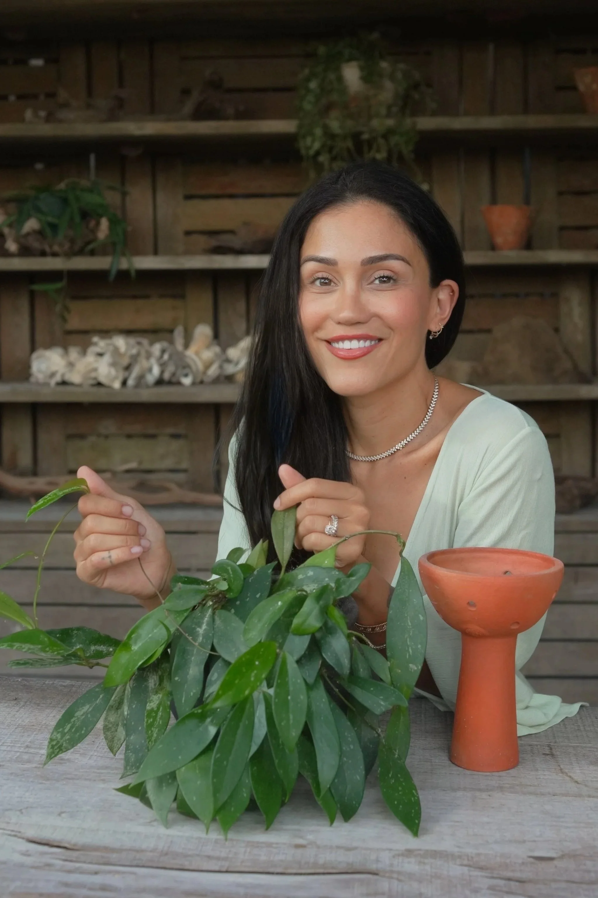 A woman with long black hair smiling at camera, sitting at a wooden table with green plants and terracotta plant holders, rustic wooden shelves with decorative items in background.