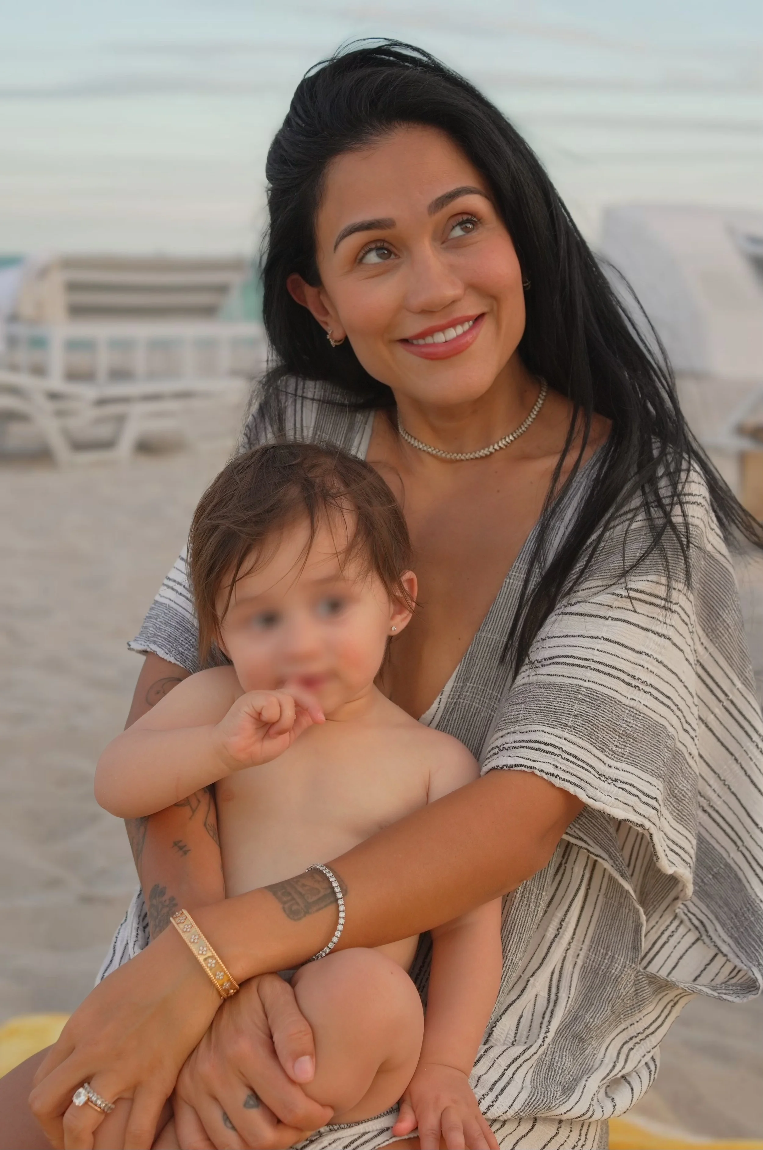 A woman with black hair, smiling, holding a young girl with wet hair at the beach, both with light to medium skin tones.
