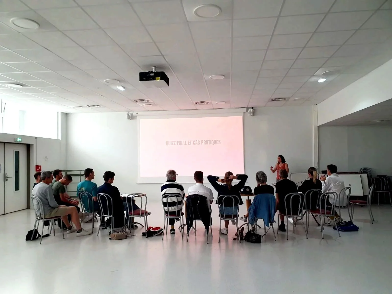 Salle de classe ou salle de conférence avec un groupe de personnes assises en cercle, une conférencière à droite, une présentation projetée à l'écran avec le titre 'Quiz final et cas pratiques'.