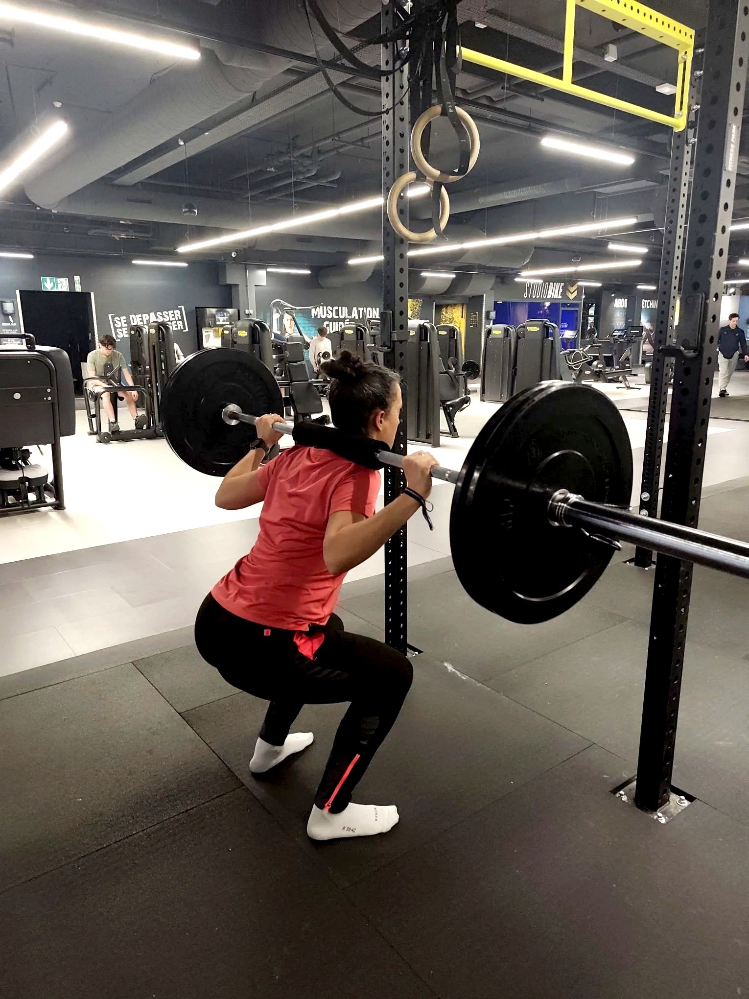 Femme en vêtements de sport faisant un squat avec une barre chargée de poids dans une salle de sport moderne.